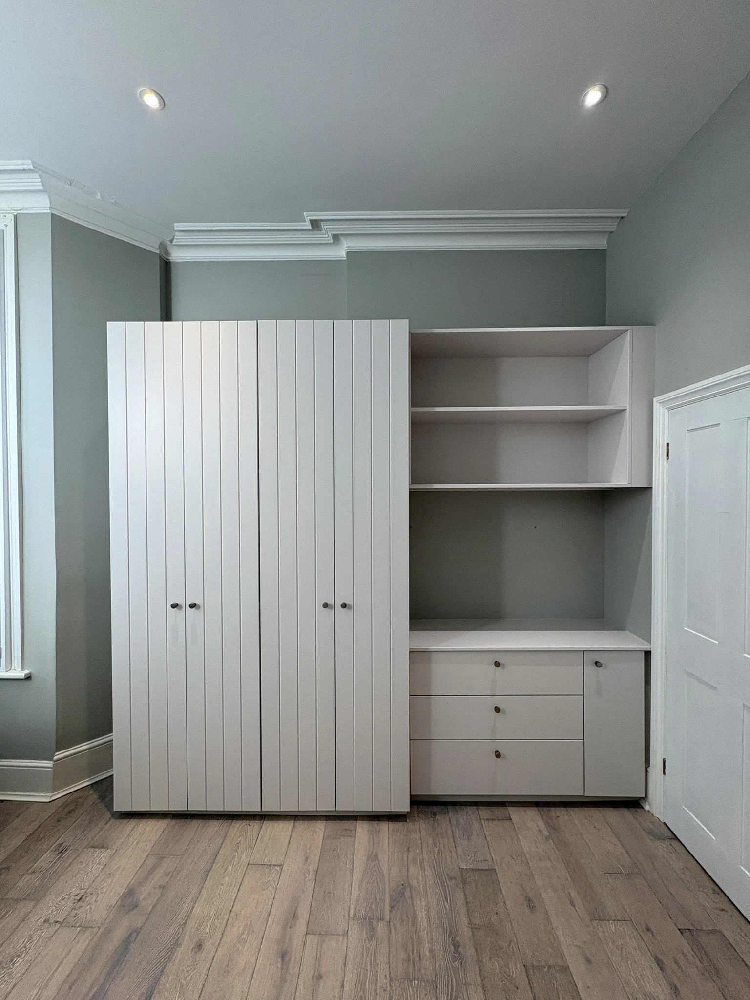 A white built-in cabinet with closed doors on the left and open shelving on the right, against a pale gray wall with crown molding and wood flooring.