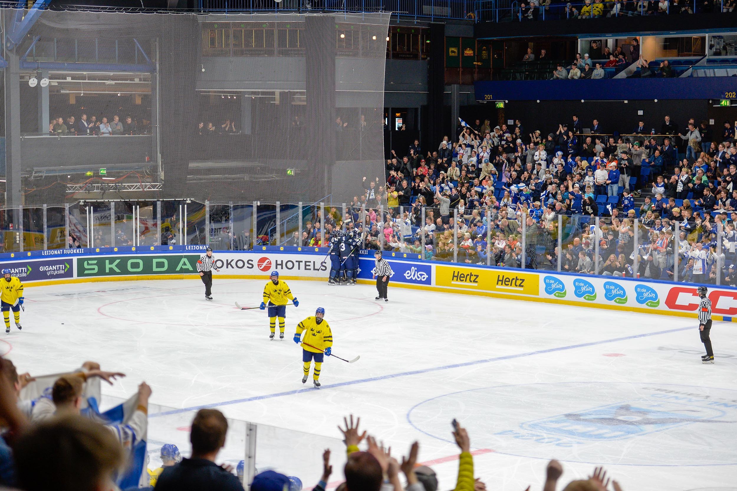Jääkiekko-ottelu, jossa yksi joukkueista pukeutunut keltaisiin peliasuihin ja toinen sinisiin. Pelaajat ovat jäällä, ja katsomossa kannattajat juhlivat. Mainoksia laitojen varrella.