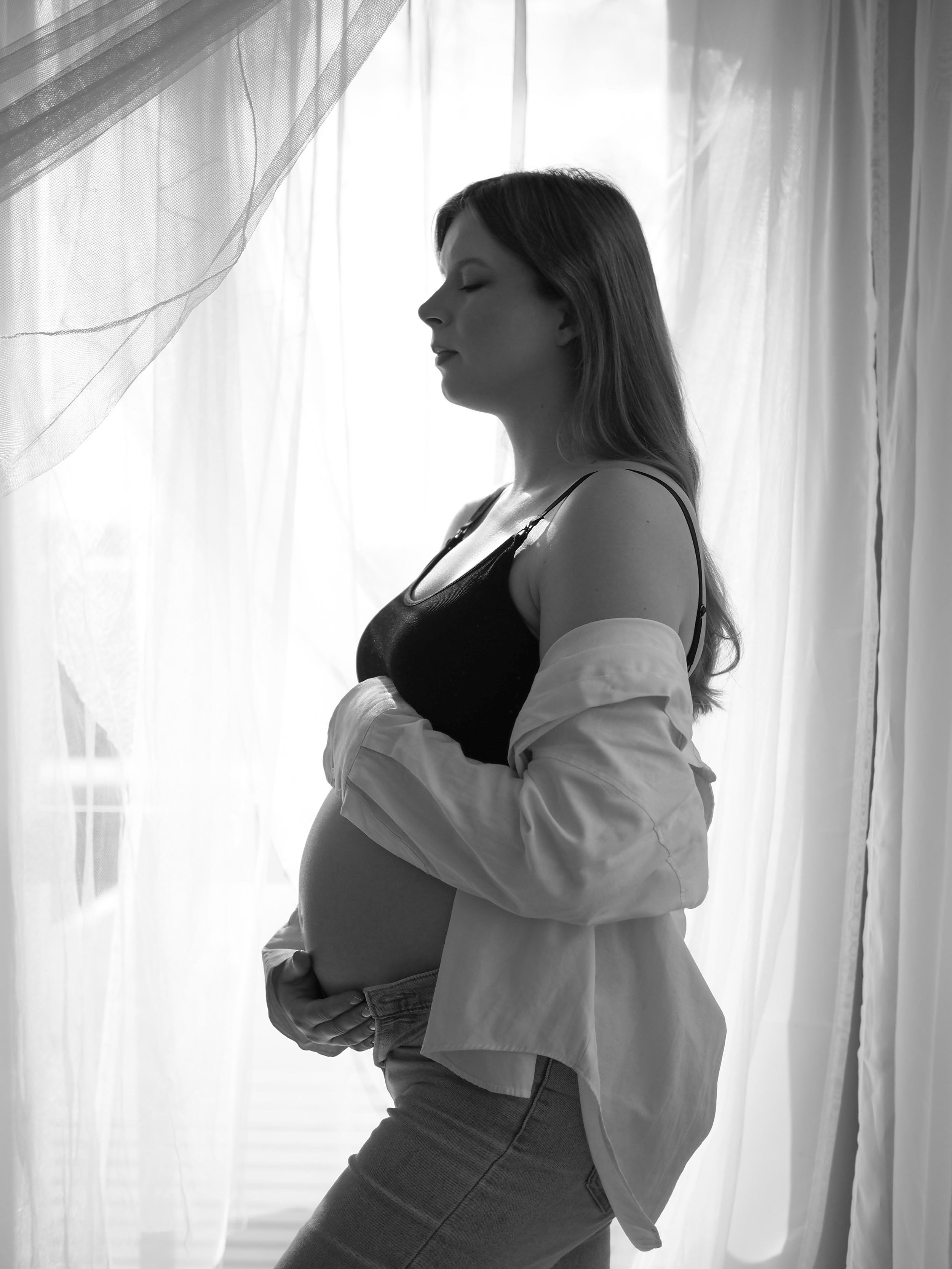 Raskaana oleva nainen profiilissa pitämässä käsiään vatsallaan, mustavalkoinen kuva verhojen edessä.