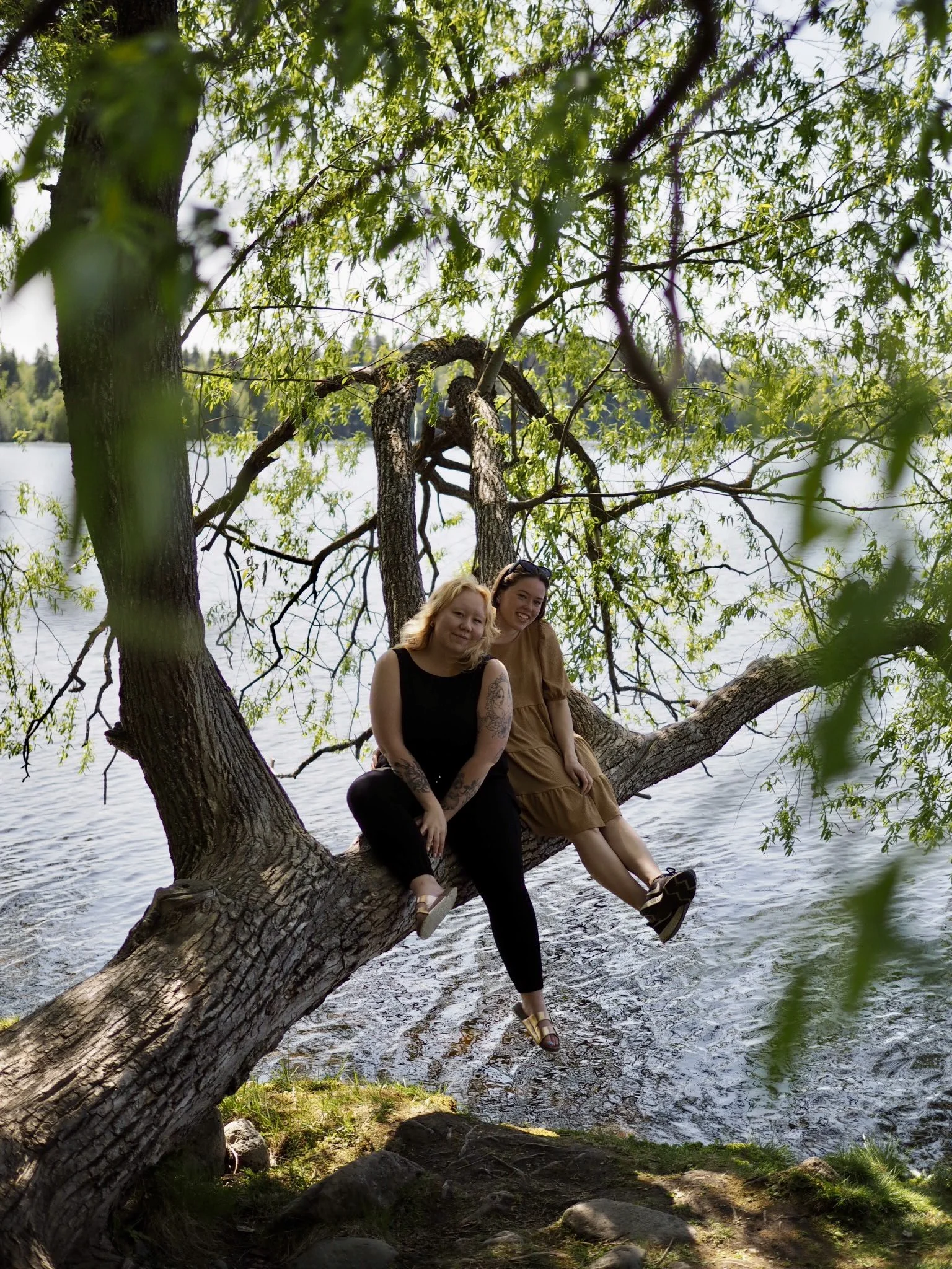 Kaksi henkilöä istuu puun oksalla veden äärellä, taustalla vehreä metsä.