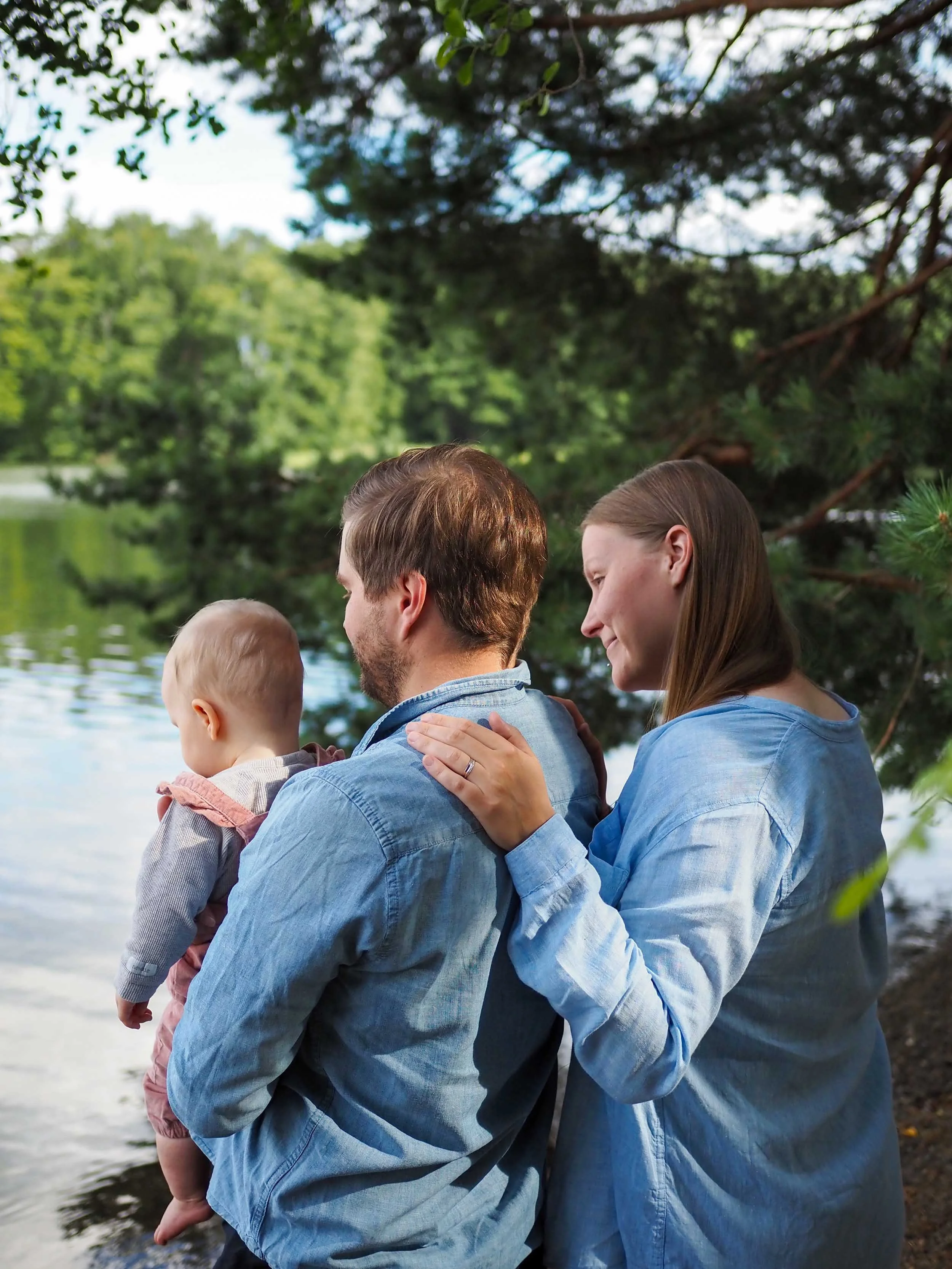 Kolme ihmistä katsoo järvelle, sylissään vauva. Taustalla on vehreitä puita.