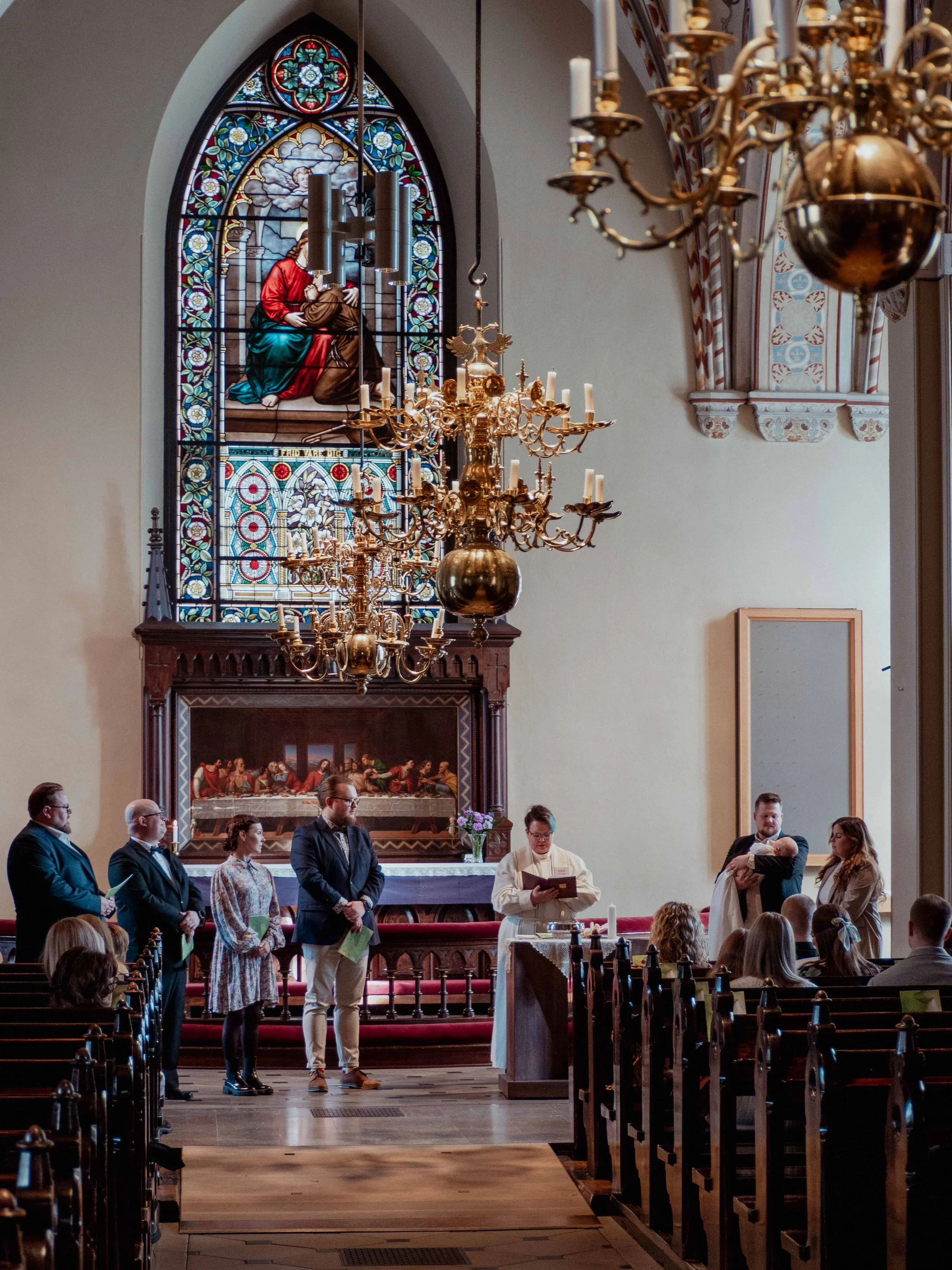 Ryhmä ihmisiä keskiaikaisessa kirkossa, jossa on värikäs lasimaalaus ja kyntteliköitä. Etualalla pappi lukee tekstiä, ja hänen ympärillään seisoo henkilöitä juhlavaatteissa. Taustalla alttaritaulu ja koristeelliset sisätilat.