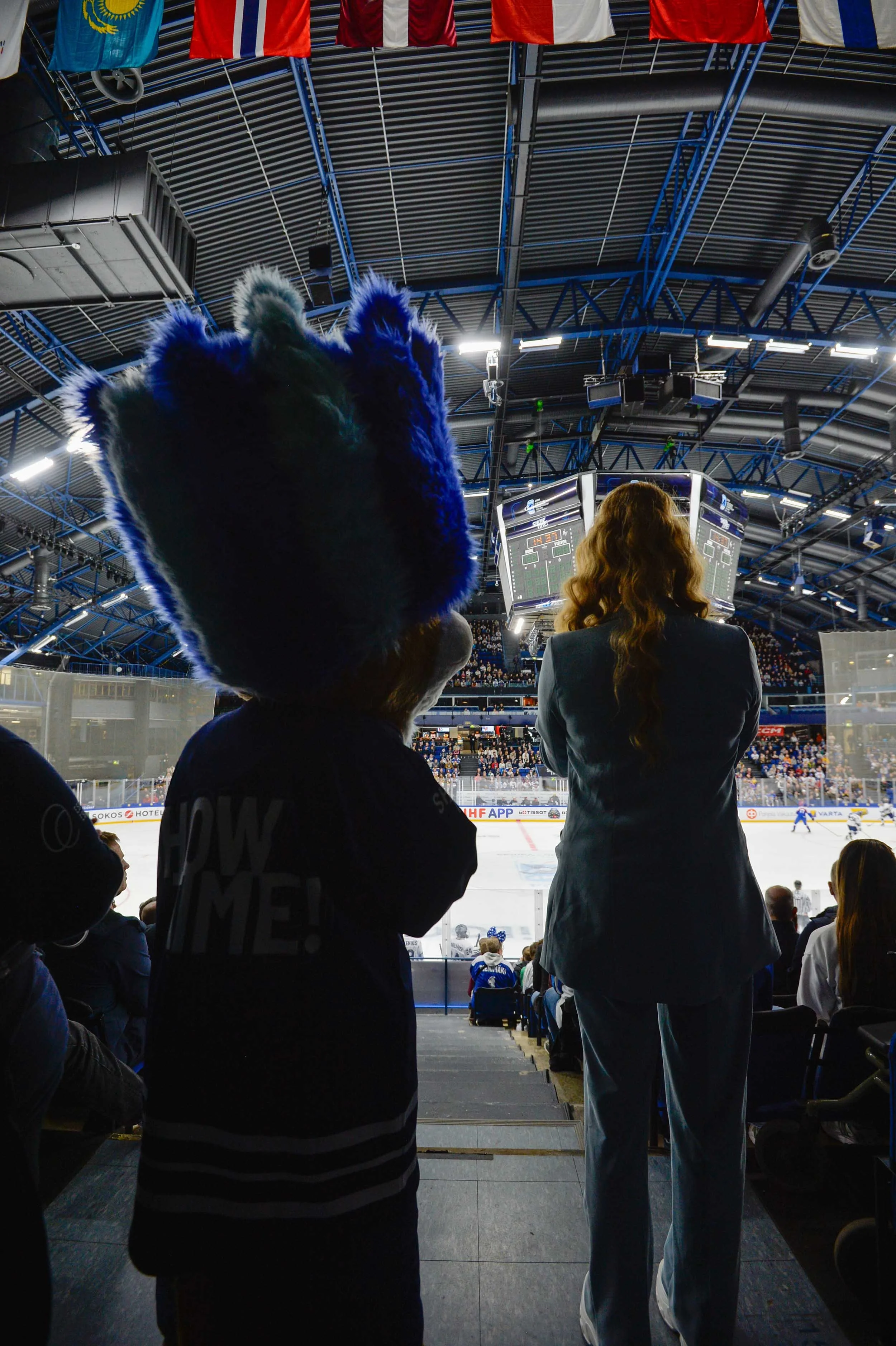 Jääkiekko-ottelu sisätiloissa, maskotti ja henkilö seisovat yleisön edessä, katsoen jääkenttää. Katossa roikkuu maiden lippuja.