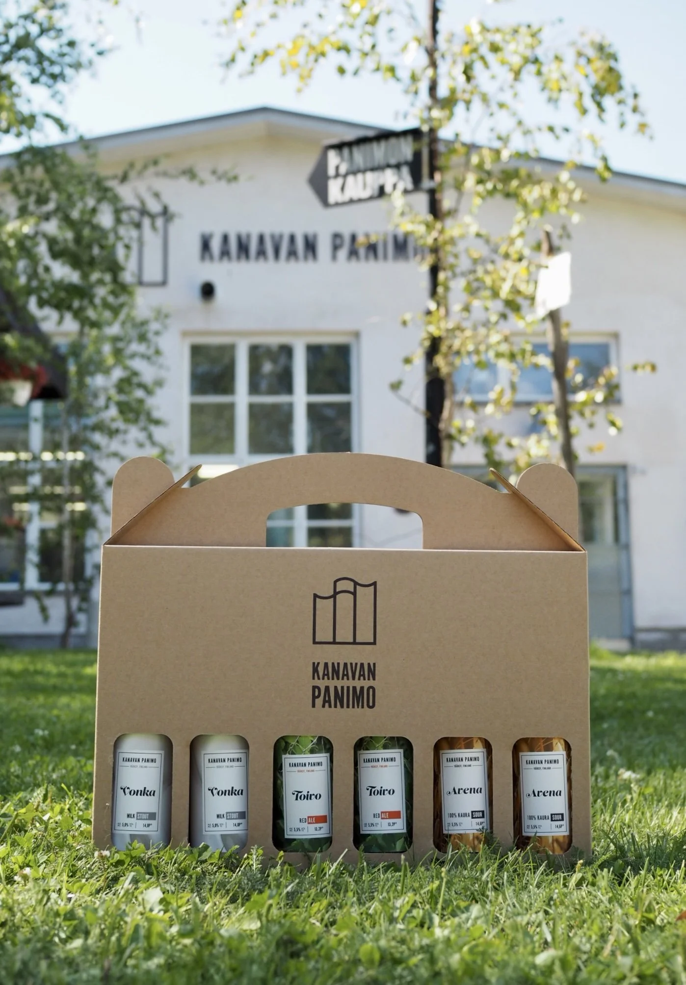Kanavan Panimon olutlaatikko, joka sisältää kuusi erilaista oluttölkkiä, rakennuksen edessä, jossa lukee "Kanavan Panimo".