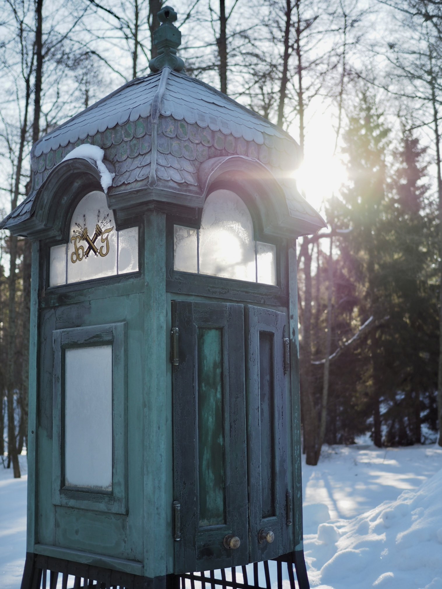 Vanha puinen puhelinkoppi talvimaisemassa, taustalla lumisia puita ja aurinko paistaa.