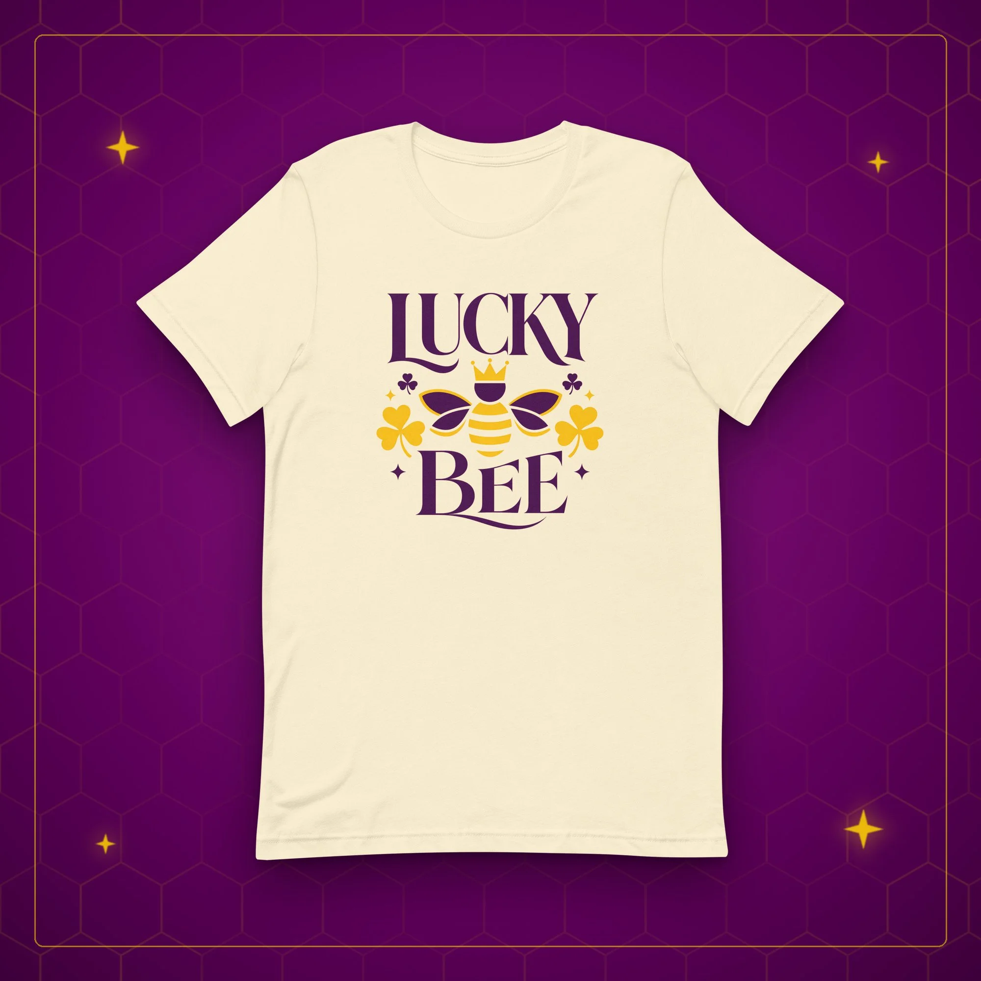 Lucky Bee tee in natural, laid flat on a purple background with gold sparkles