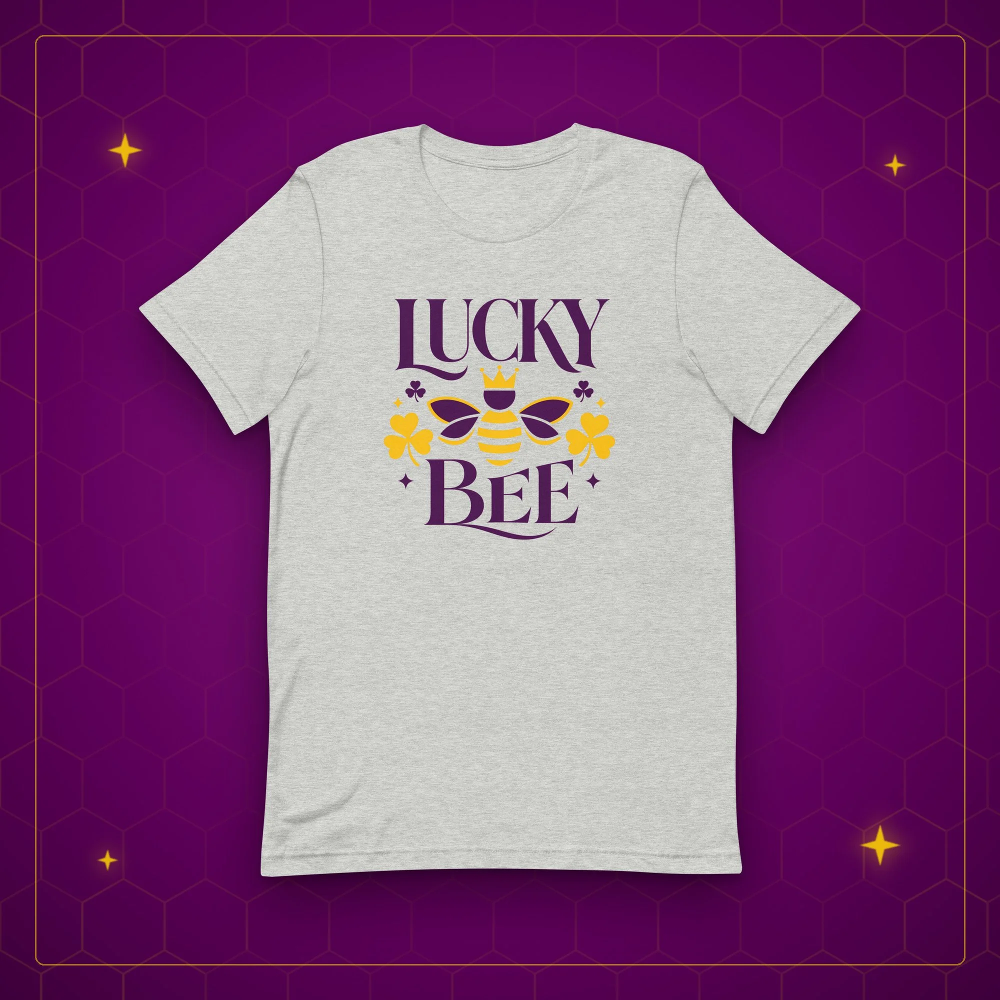 Lucky Bee tee in athletic heather, laid flat on a purple background with gold sparkles