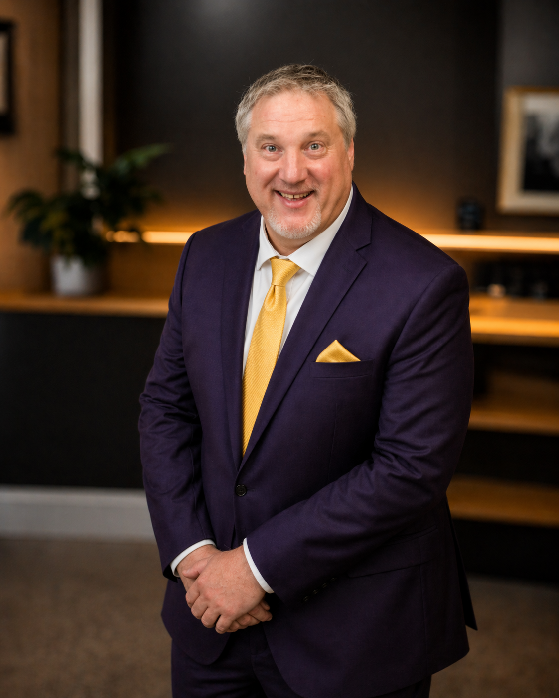 A smiling man in a dark suit with a yellow tie and pocket square, standing in a modern office or conference room with wood and black accents, and a potted plant in the background.
