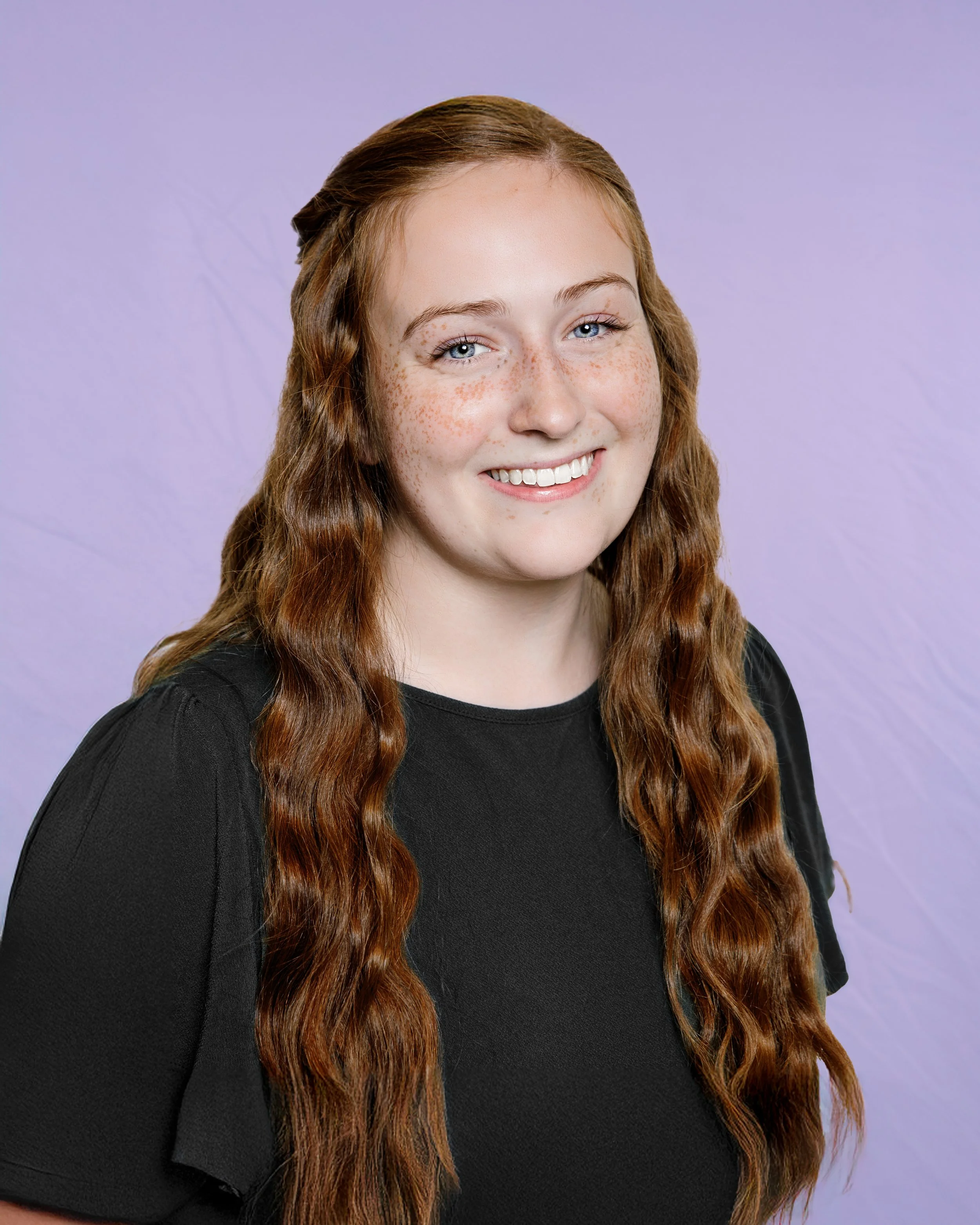 Portrait of a young woman with long red curly hair, freckles, smiling, wearing a dark green top.