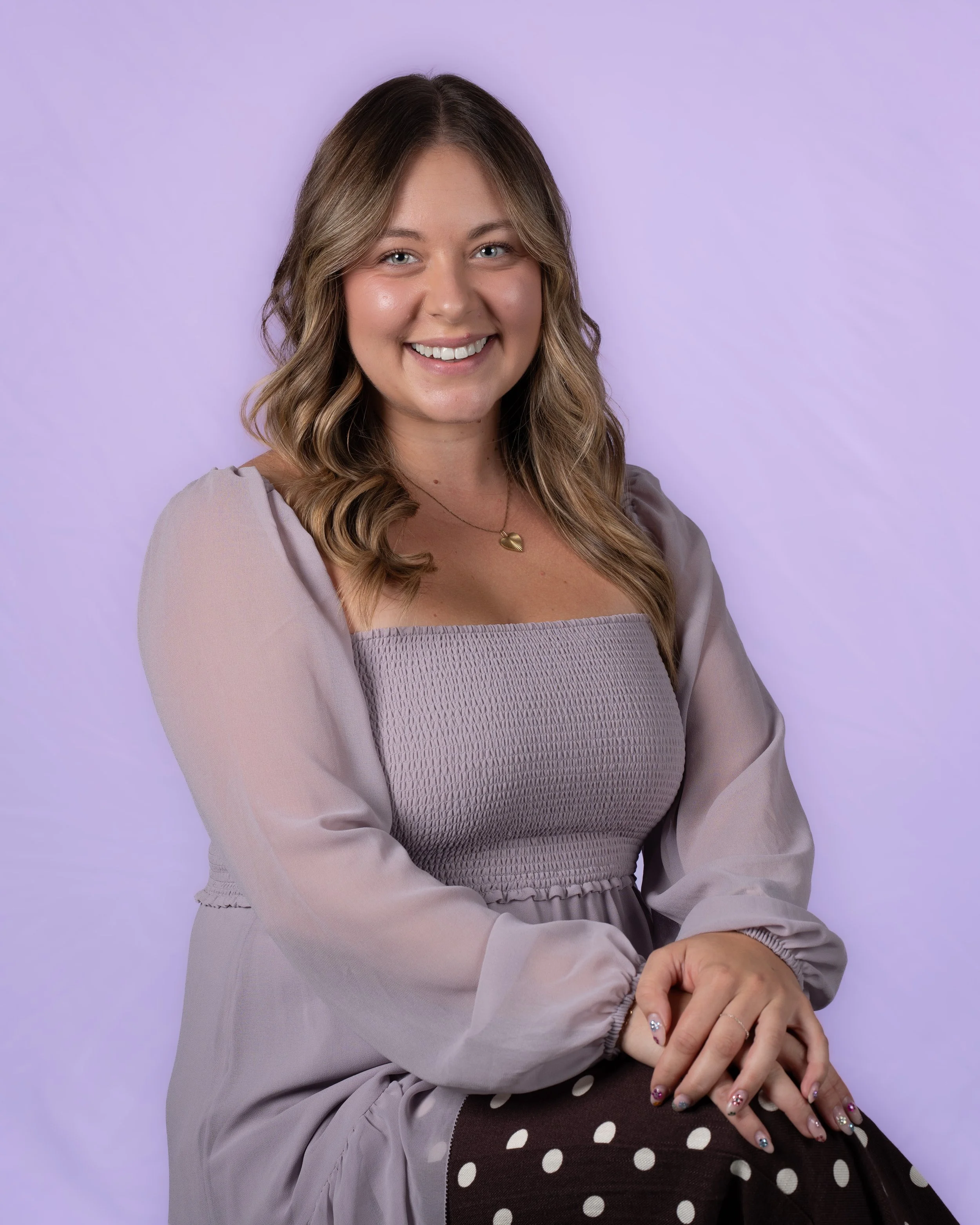 Close-up portrait of a young woman with light brown hair, blue eyes, and fair skin smiling, wearing a sleeveless gray top and small hoop earrings, standing against a plain white background.
