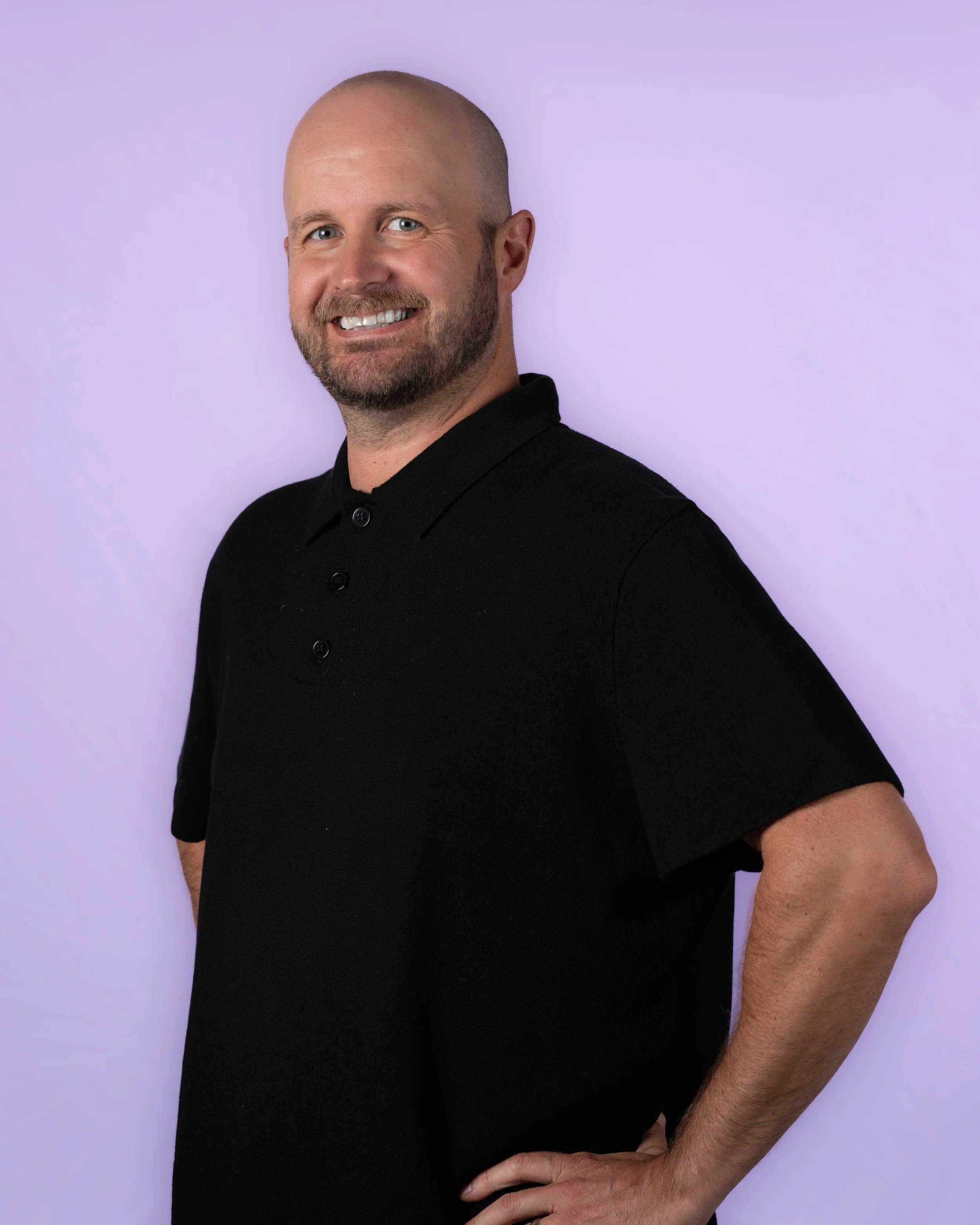 A man with a beard and no hair smiling, wearing a black shirt with a plaid collar and folded arms, against a gray background.