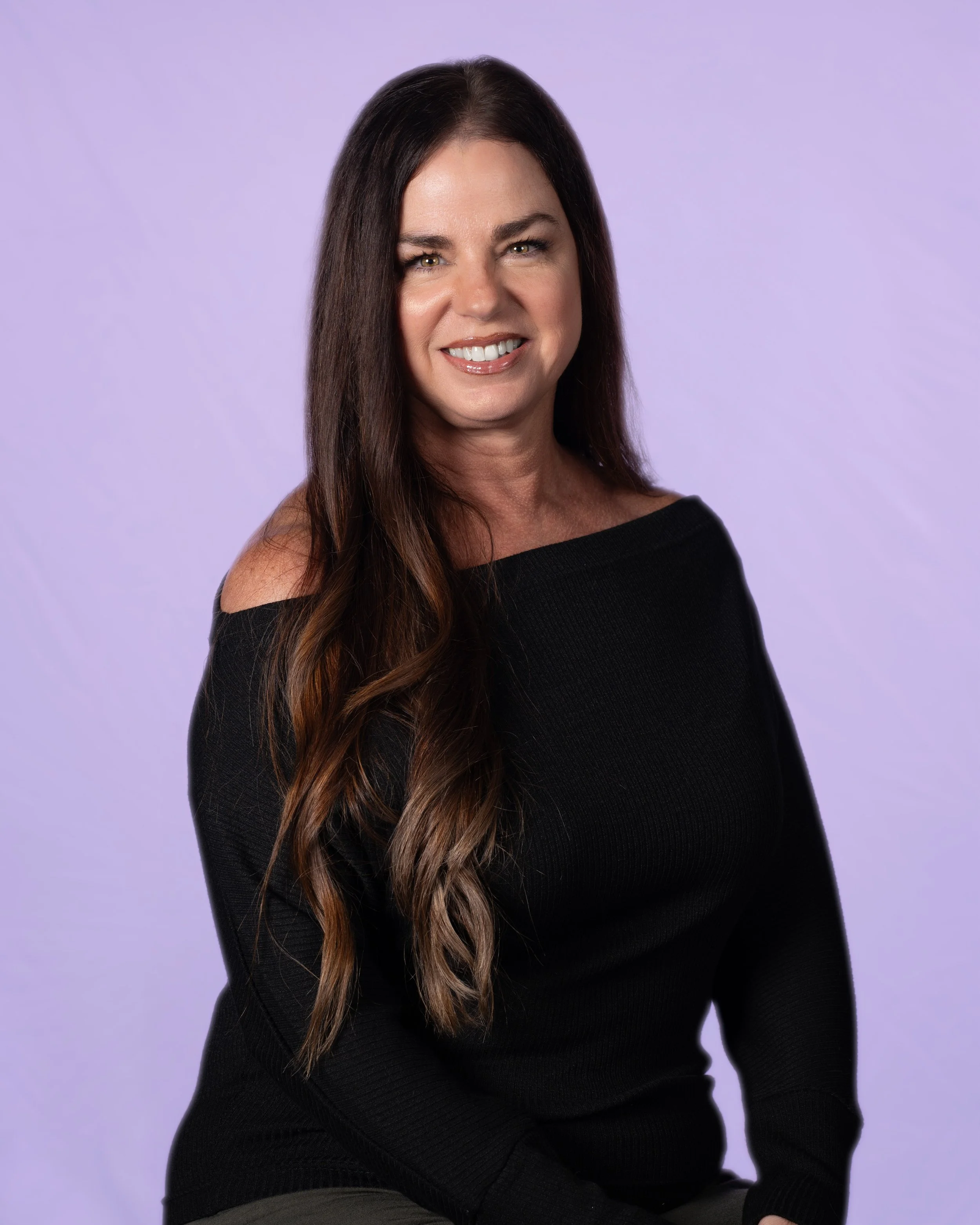 A woman with long dark hair wearing a light gray off-shoulder top smiling at the camera.