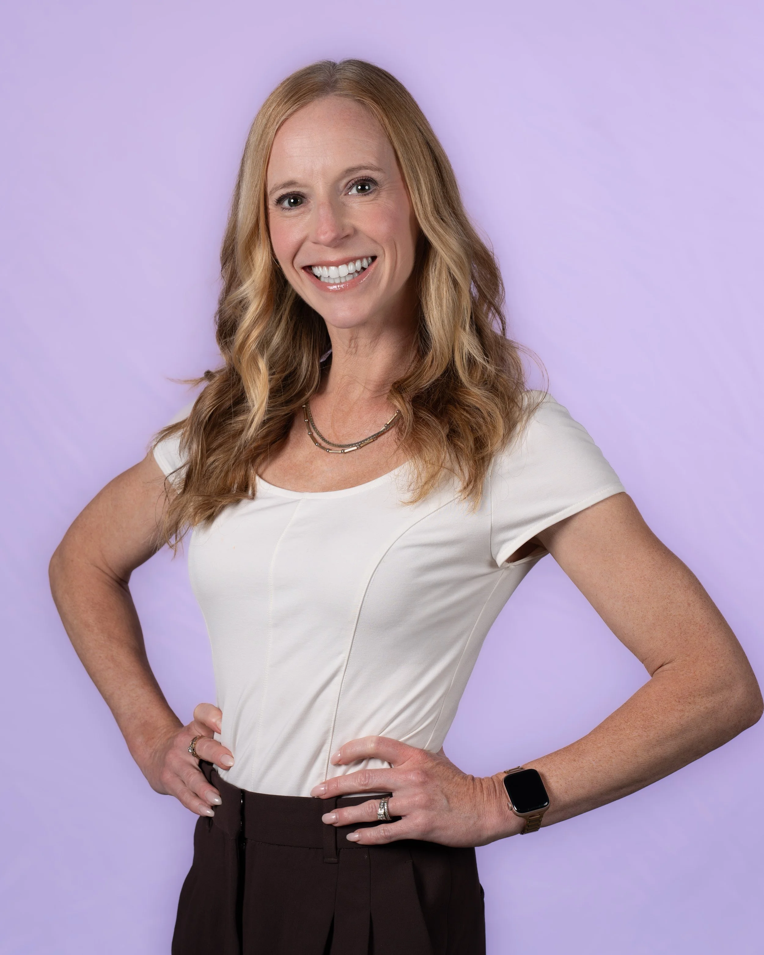 A woman with shoulder-length reddish-blonde hair, smiling, wearing a black top against a gray background.