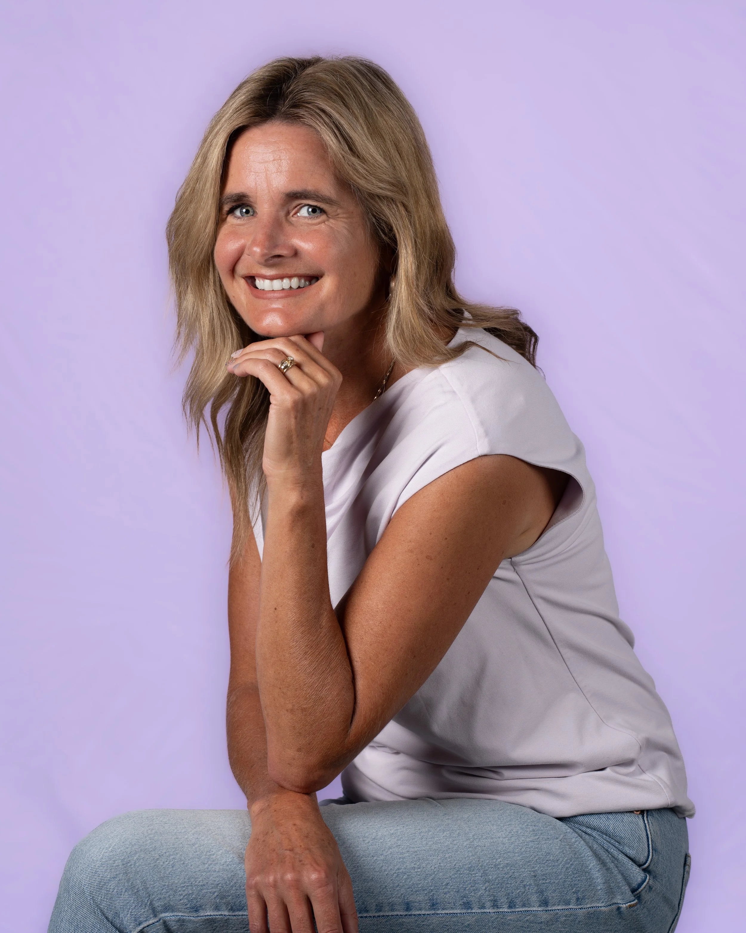 A woman with shoulder-length light brown hair smiling, wearing a white button-up shirt with chest pockets, and dark pants, standing against a white background.