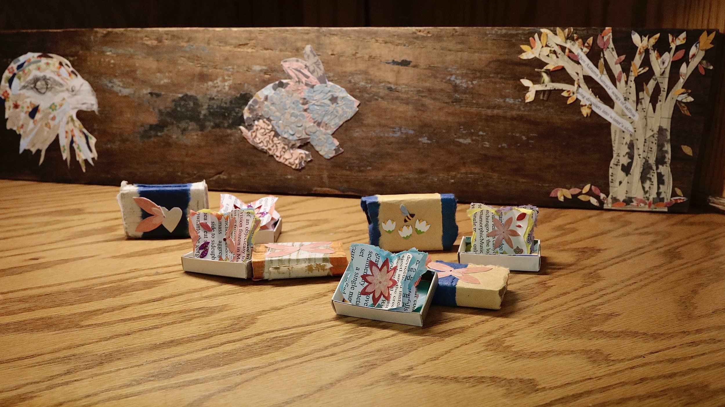 Wooden board with paper collage art featuring a tree, rabbit, and bird; small boxes with decorative paper cutouts on a wooden surface.