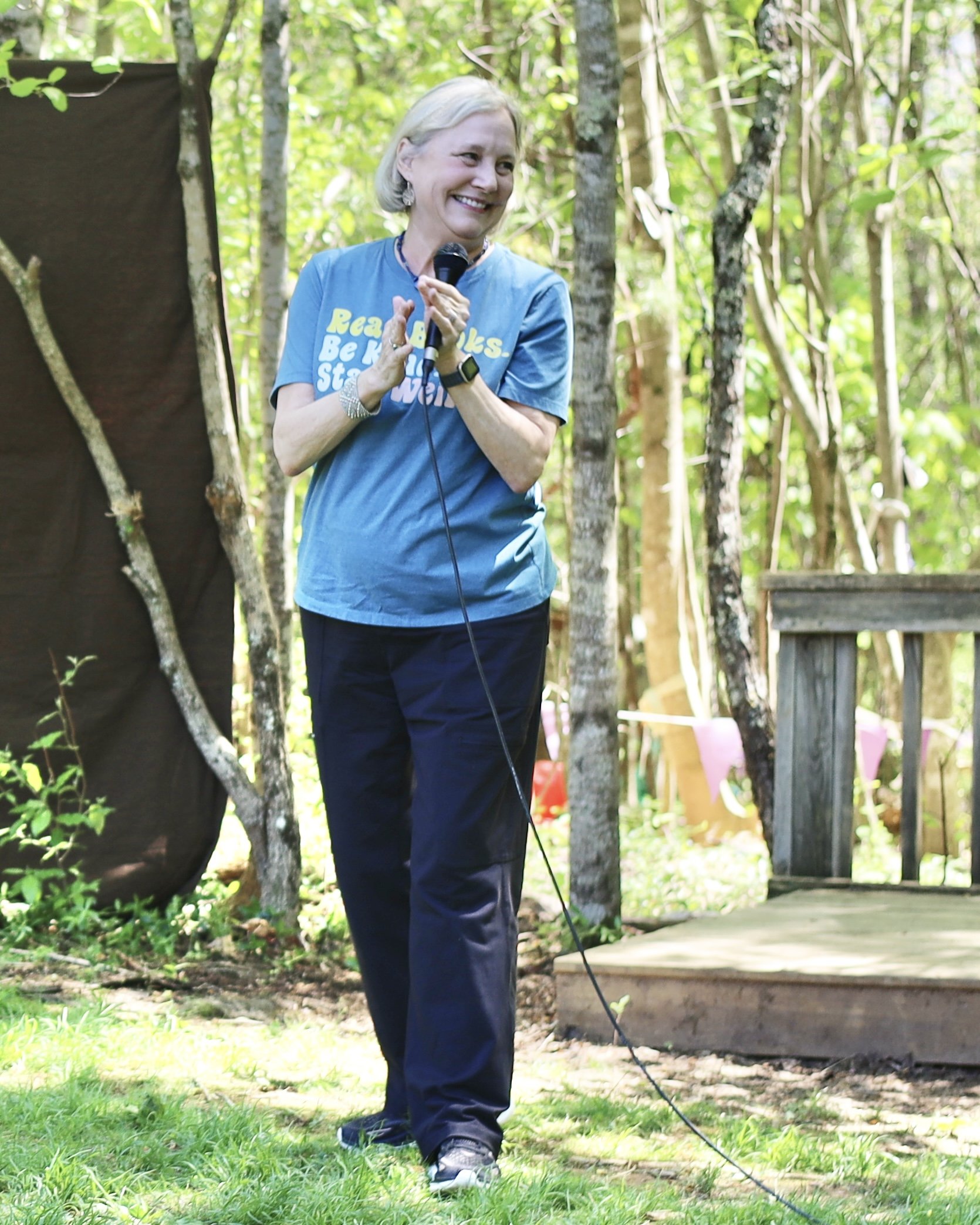 A person standing outdoors with a microphone, wearing a blue T-shirt with text, surrounded by trees and greenery.