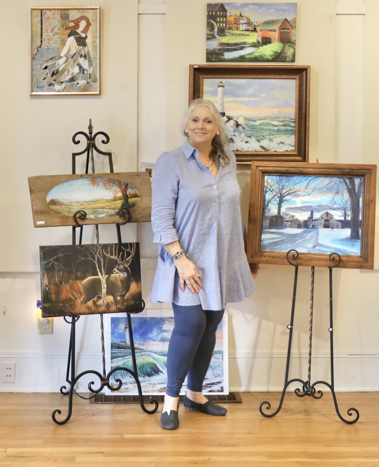 Person standing in front of easels displaying paintings, including landscapes and a winter scene, in an art gallery.
