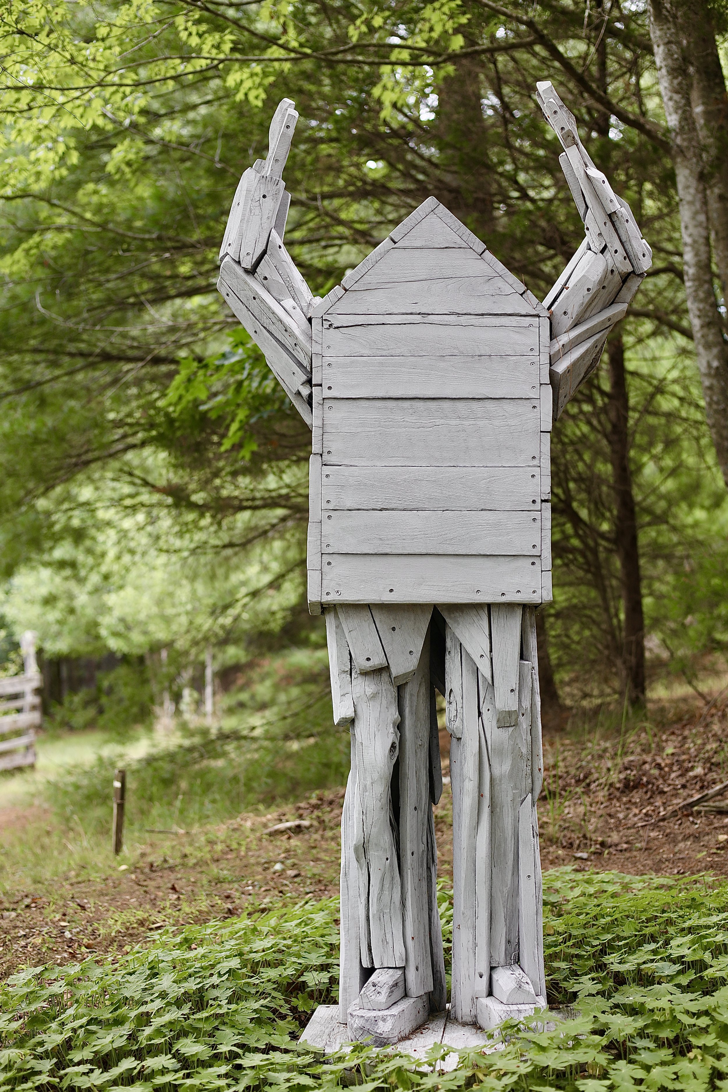 Wooden humanoid sculpture with raised arms in a forest setting