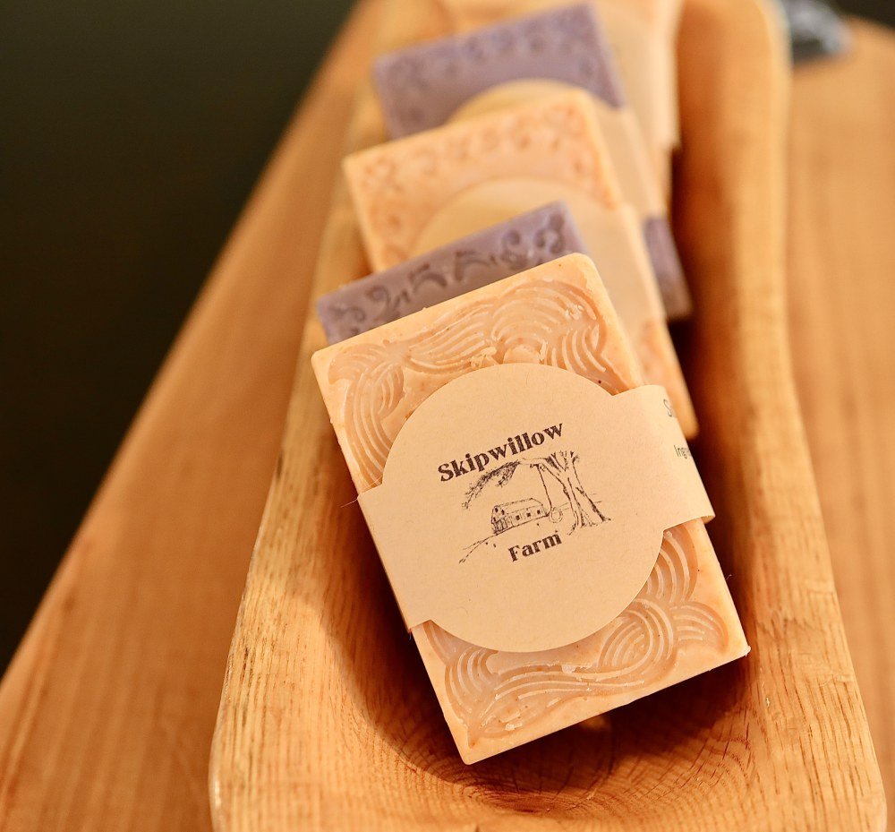 Handmade soap bars with decorative patterns, labeled "Skipwillow Farm," arranged in a wooden tray.