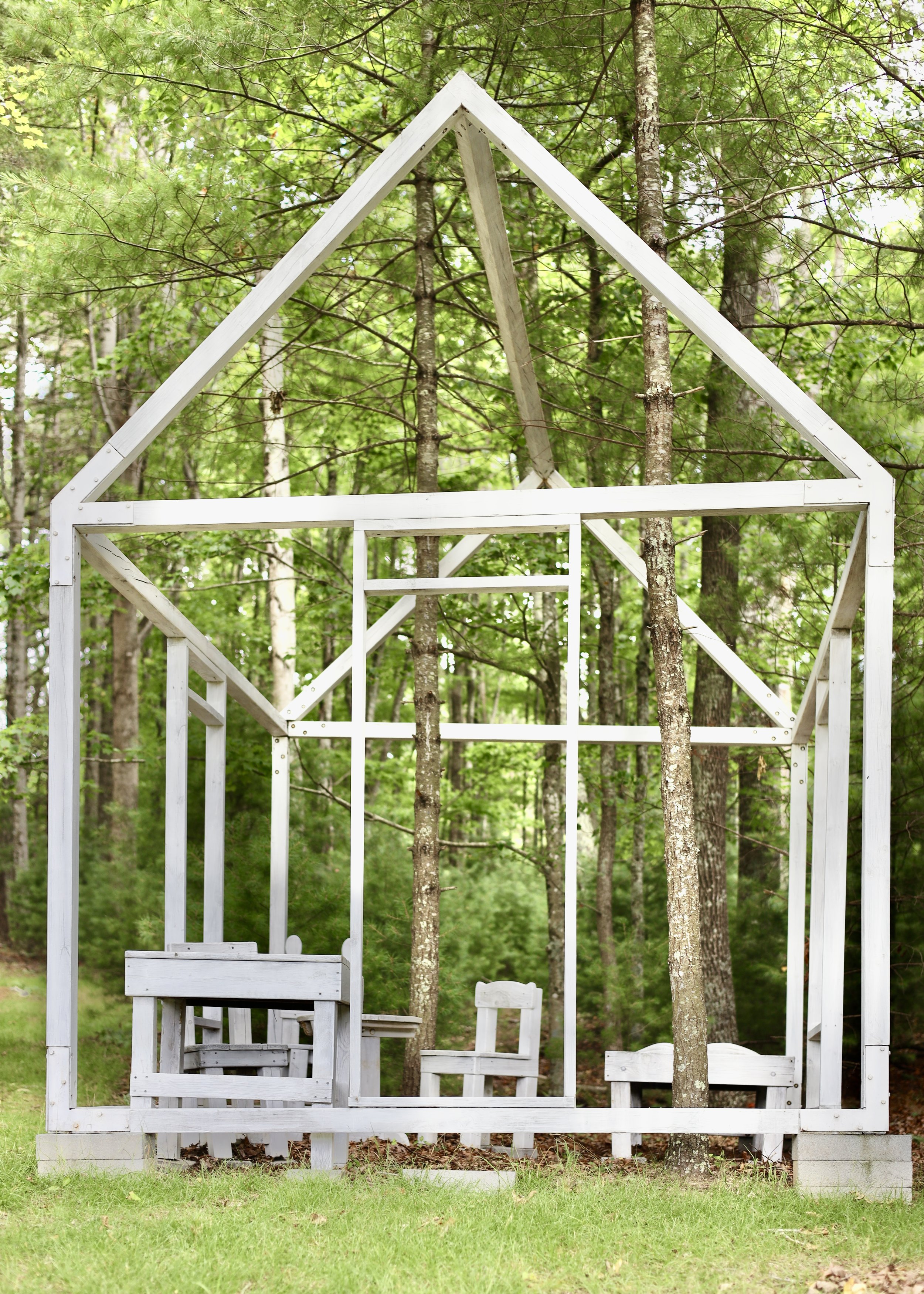 White wooden frame of a house structure in a forest, with trees growing through it. It includes a table and chairs made of the same material.