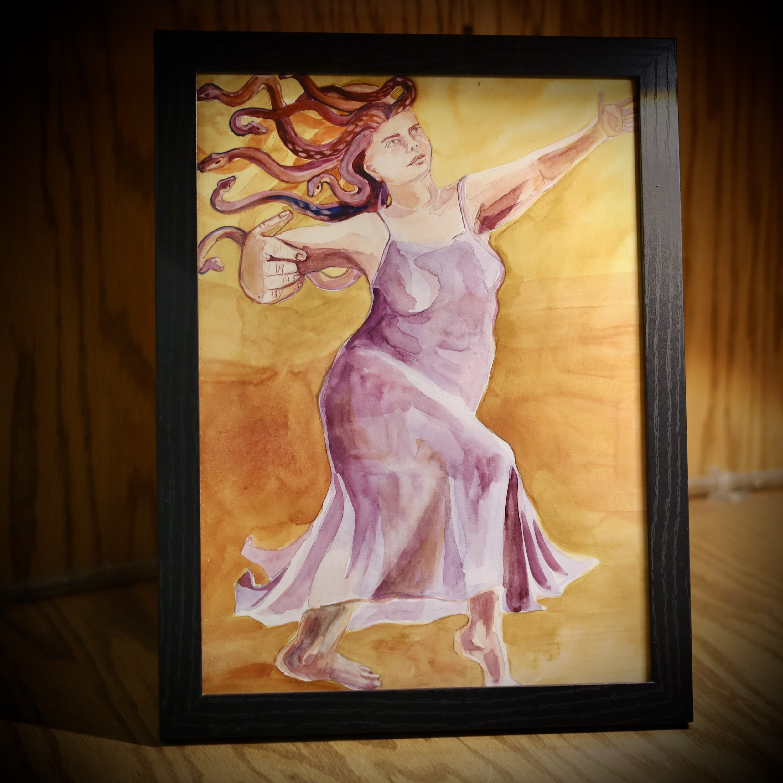 Framed watercolor painting depicting a woman dancing. She is wearing a flowing purple dress with her long hair swirling around, set against a warm orange background.
