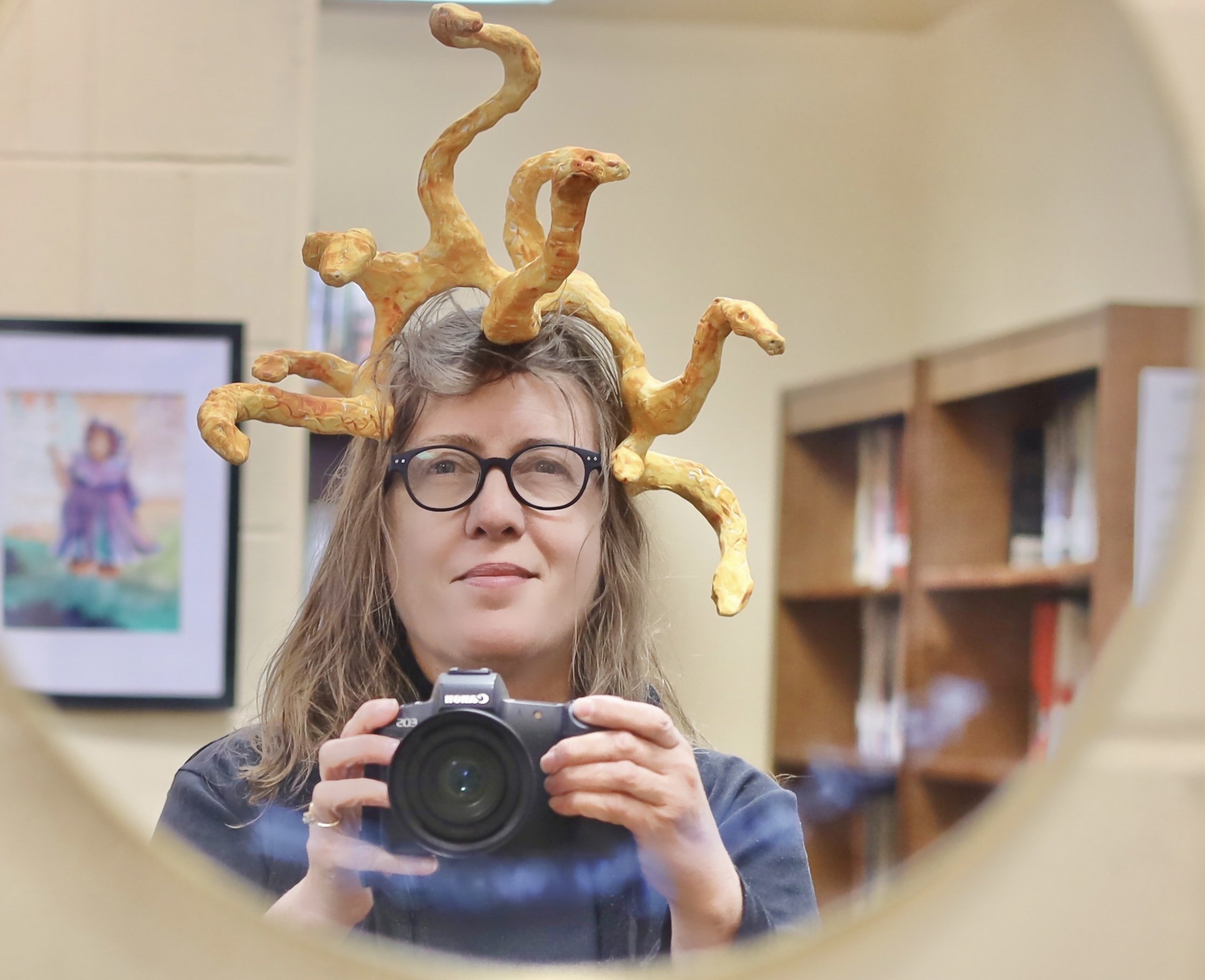 Person with glasses holding a camera, wearing a Medusa-like headpiece with snake-like extensions, in an indoor setting with shelves and a framed picture in the background.