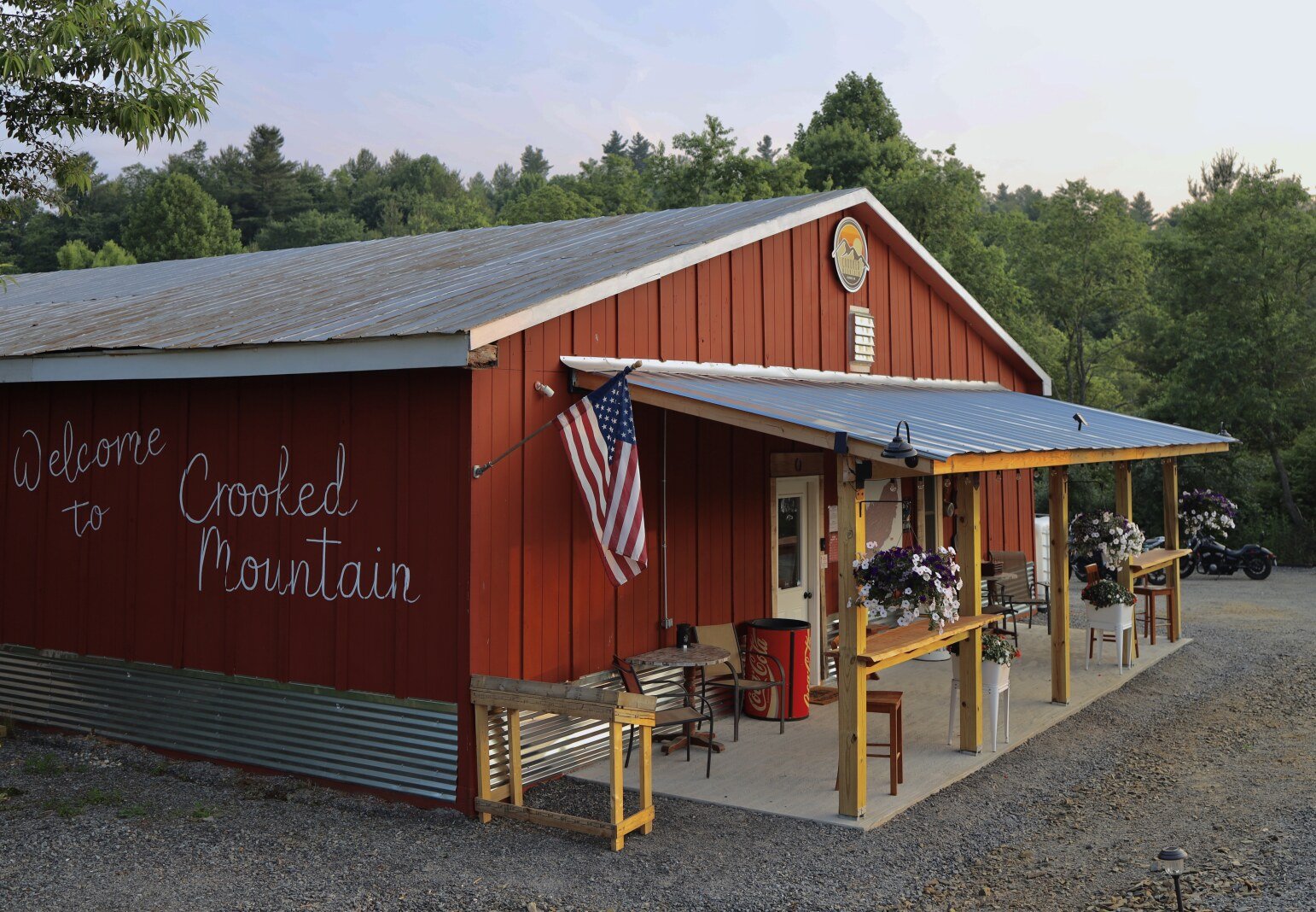 Red metal building with "Welcome to Crooked Mountain" written on the side, an American flag hanging, outdoor seating, potted flowers, and wooded background.