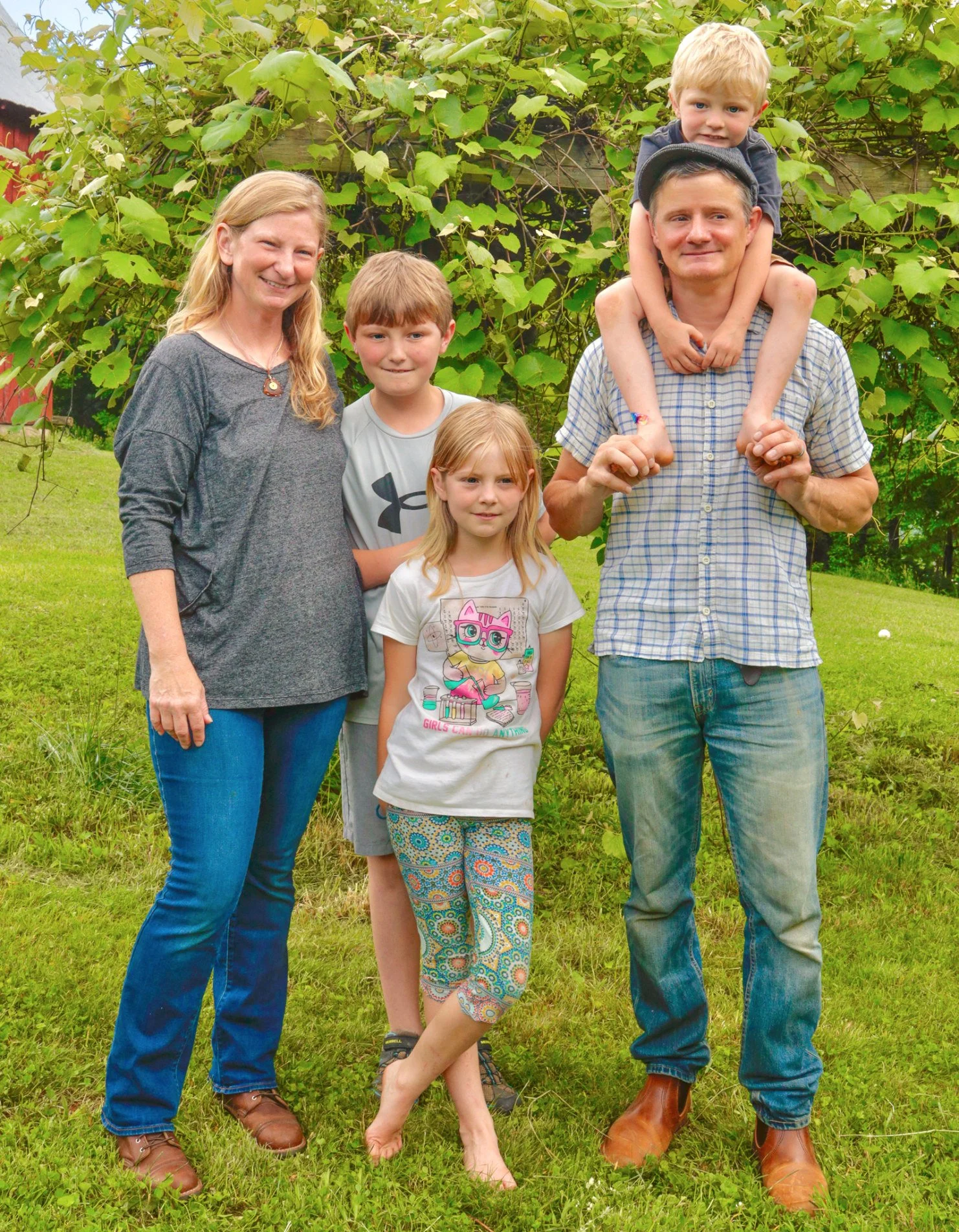 A family of six standing in a grassy area with a leafy background. The father is wearing a checkered shirt and jeans, holding a young child on his shoulders. The mother is wearing a gray top and jeans. Two children are standing in front of the parent