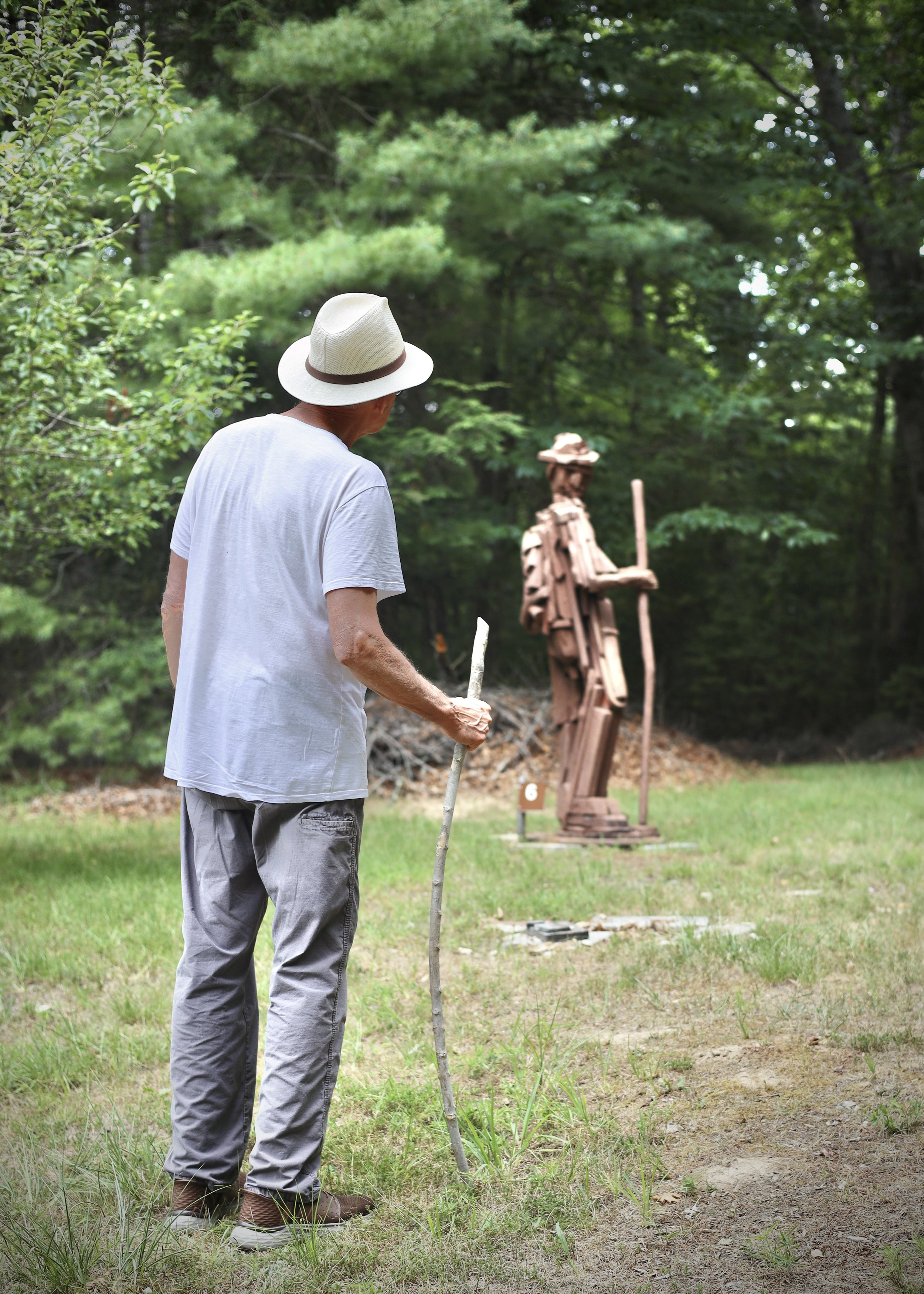 A person wearing a hat and holding a stick stands on grass, observing a wooden sculpture in a forested area.