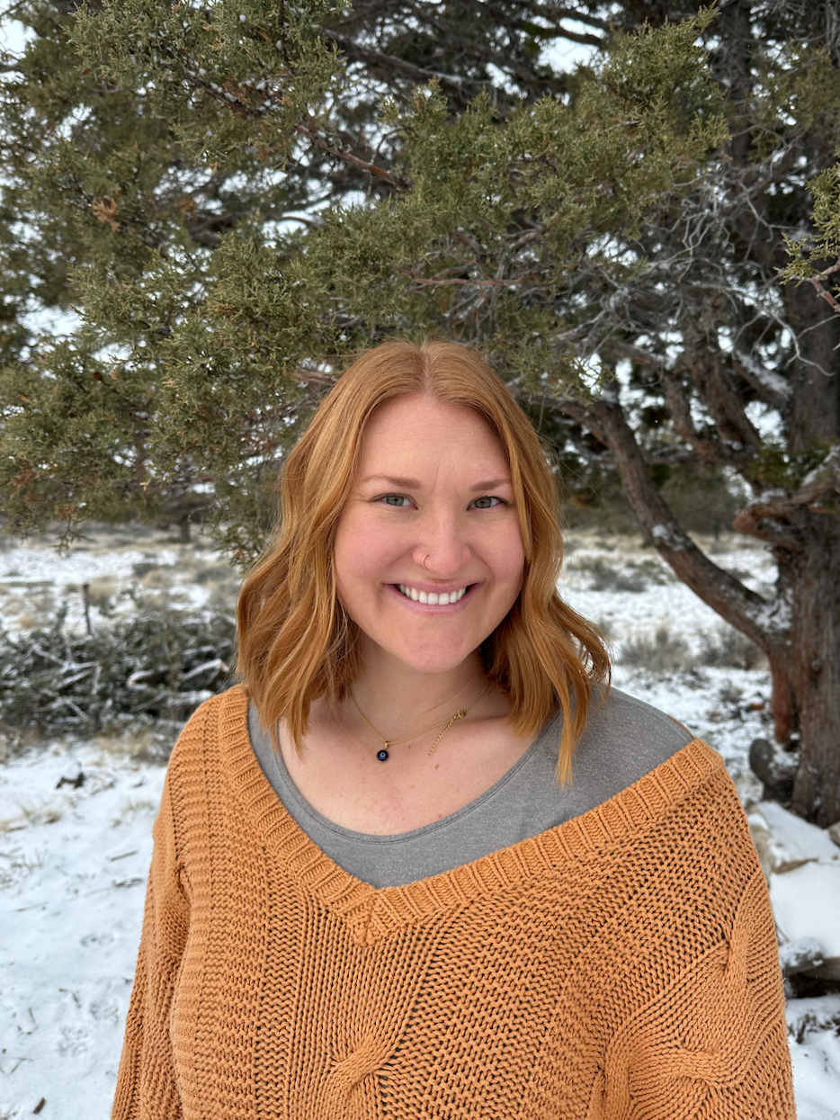 A woman with red hair smiling outdoors in winter, standing in front of a pine tree with snow on the ground.