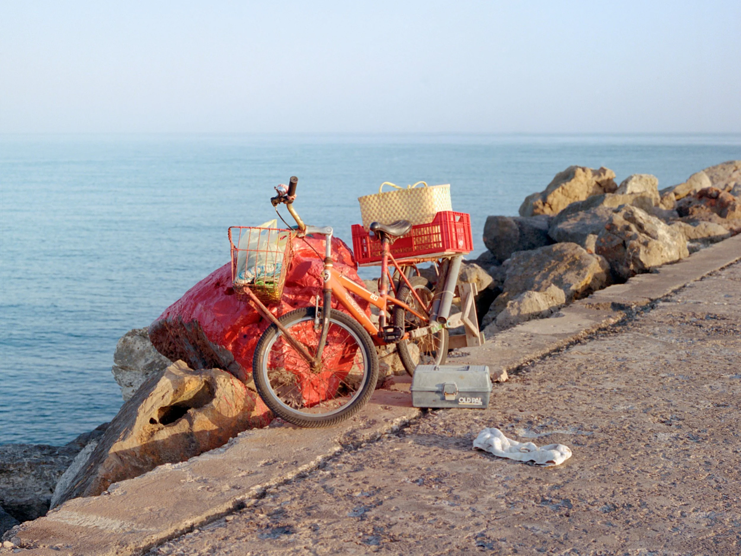 Bicycle with basket and crate parked on a rocky pier by the sea; tackle box and cloth nearby.