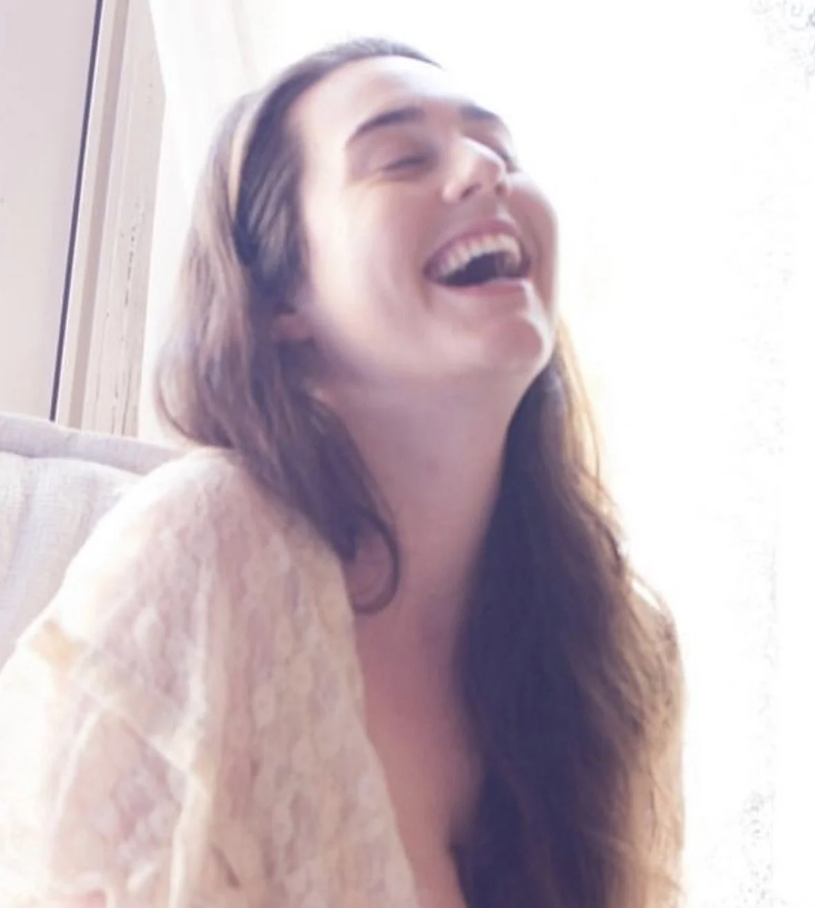 A woman with long brown hair laughing with her eyes closed, sitting on a sofa near a window.