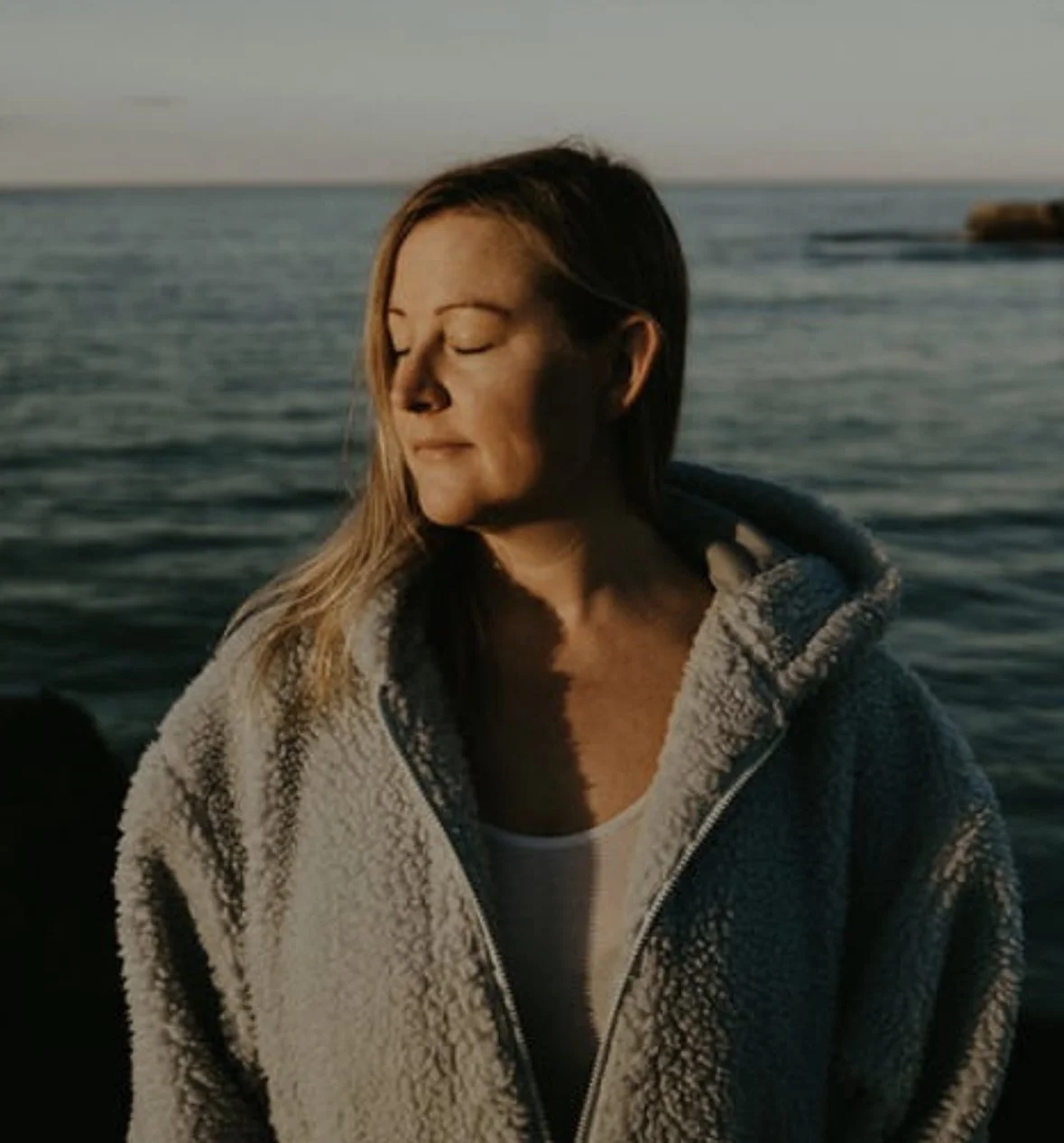 A woman with closed eyes and a relaxed expression stands near the water during sunset, wearing a gray fleece jacket.