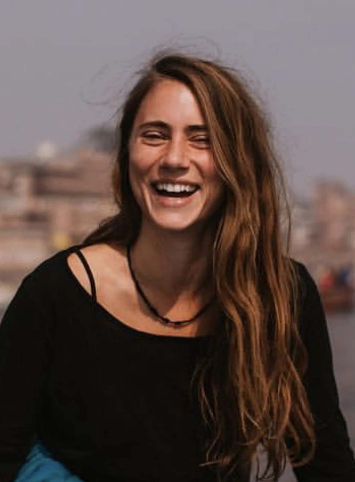 A woman with long red hair, smiling and laughing, outdoors with blurred cityscape in the background.