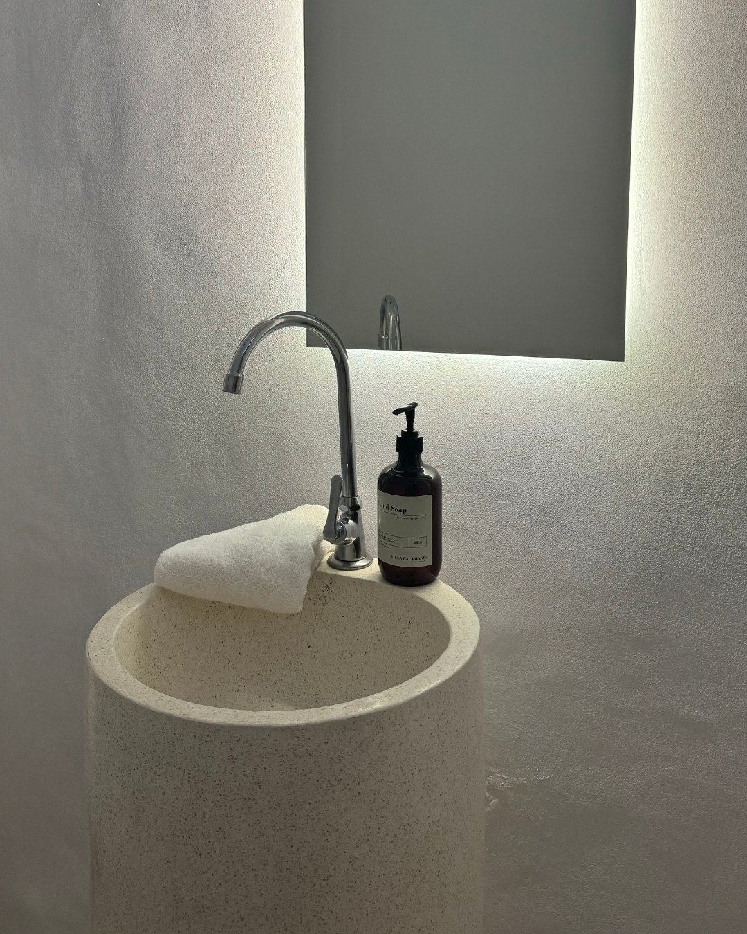 Minimalist stone sink with silver tap withhand wash in an amber glass pump bottle and white folded towel on the back of the sink. A backlit mirror is on the neutral wall behind.