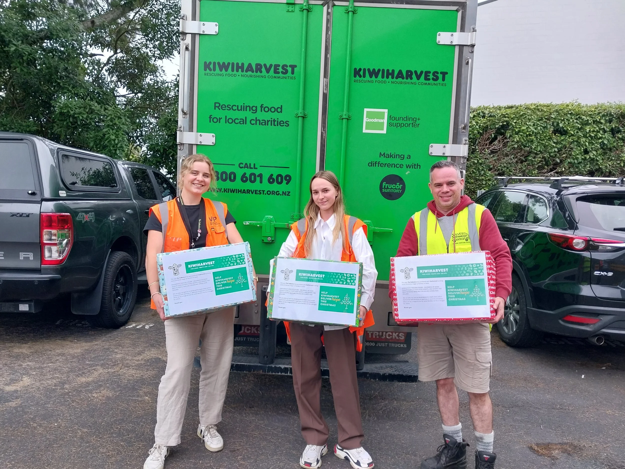 Three volunteers stand behind the KiwiHarvest food rescue truck, holding food donation boxes