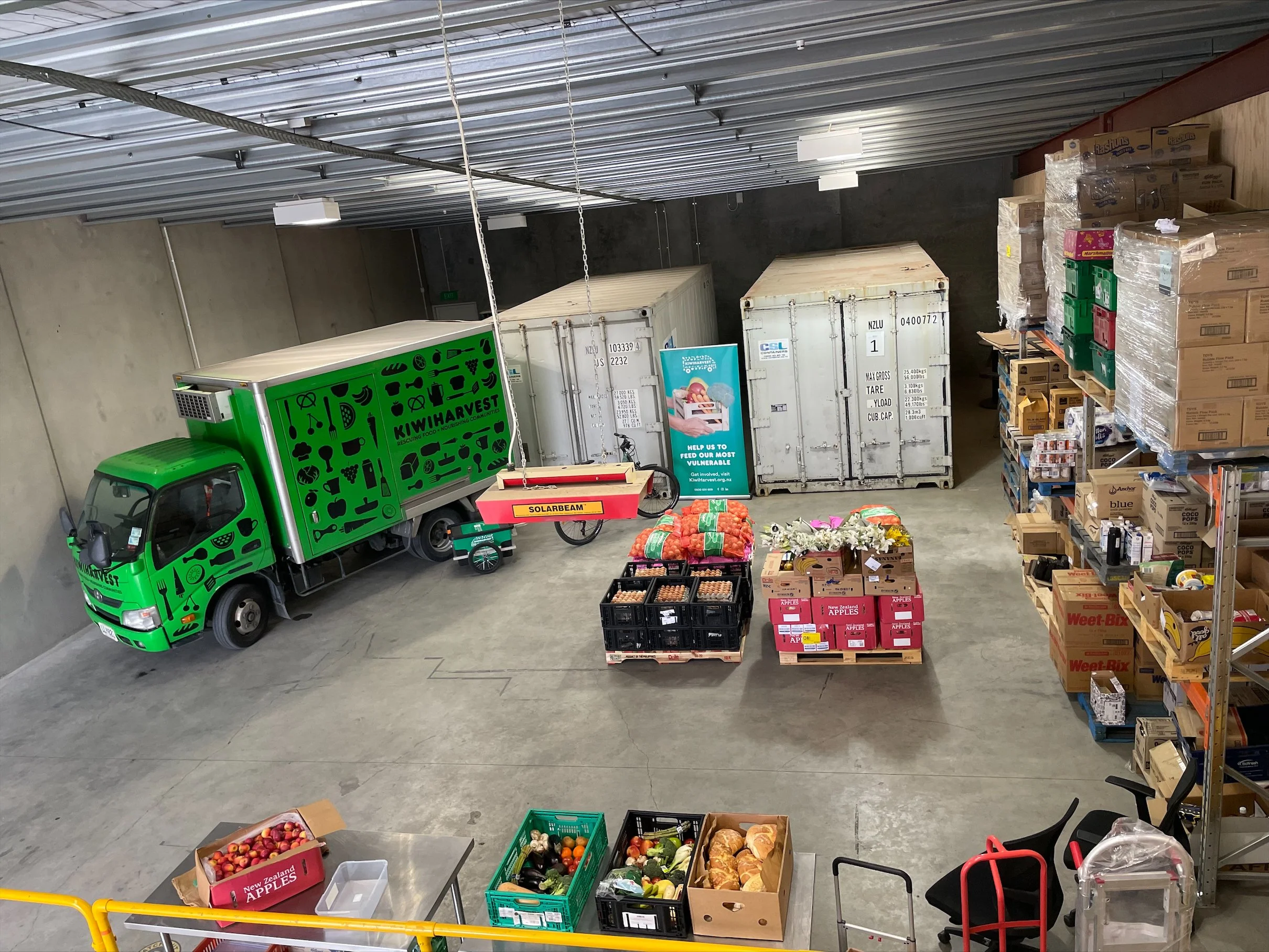 Long shot of KiwiHarvest Queenstown warhouse with fresh food sitting in boxes, ontop of crates. The Green KiwiHarvest truck sits parked in the warehouse.