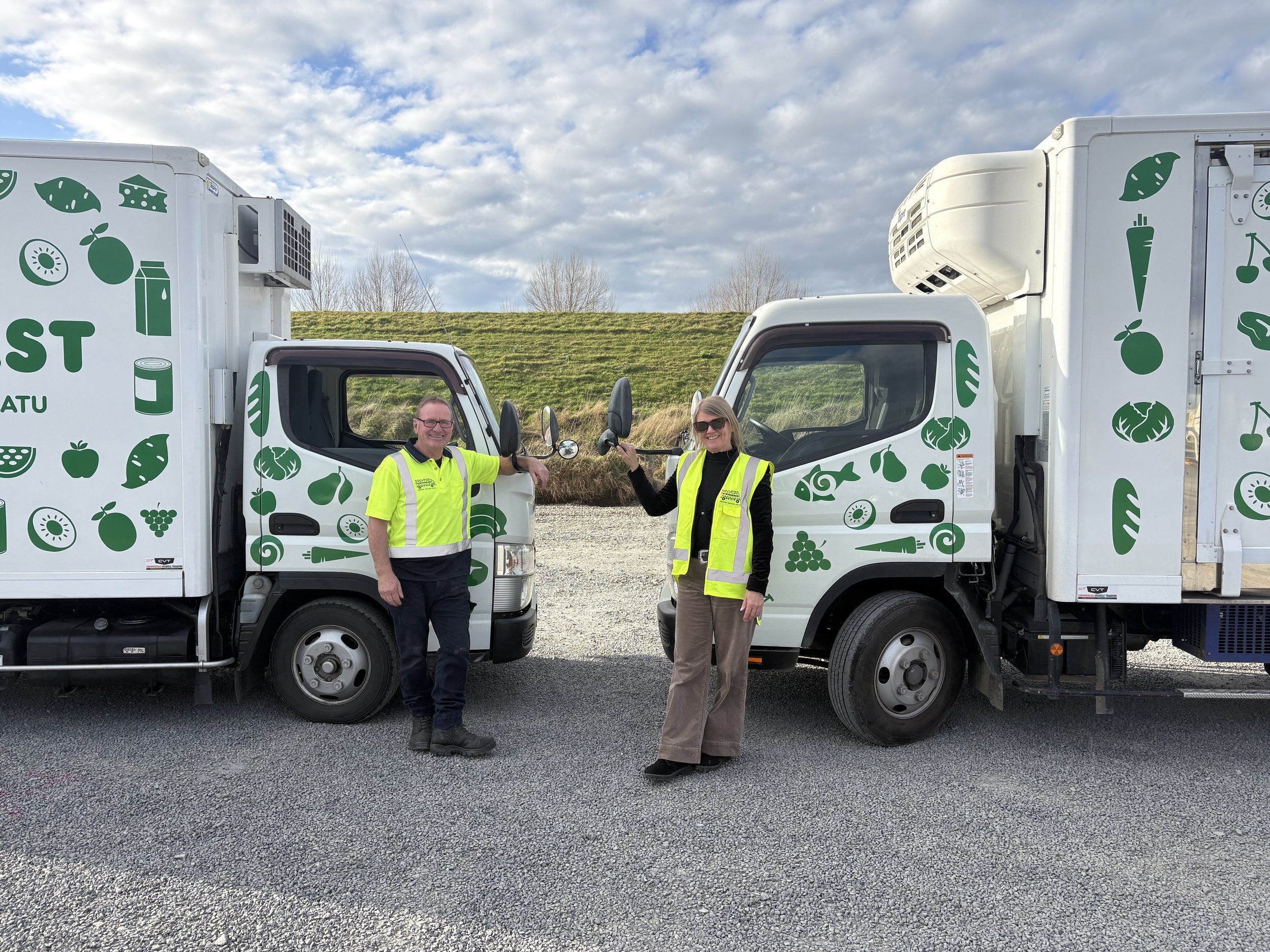 two KiwiHarvest team members stand side by side smiling infront of their KiwiHarvest trucks