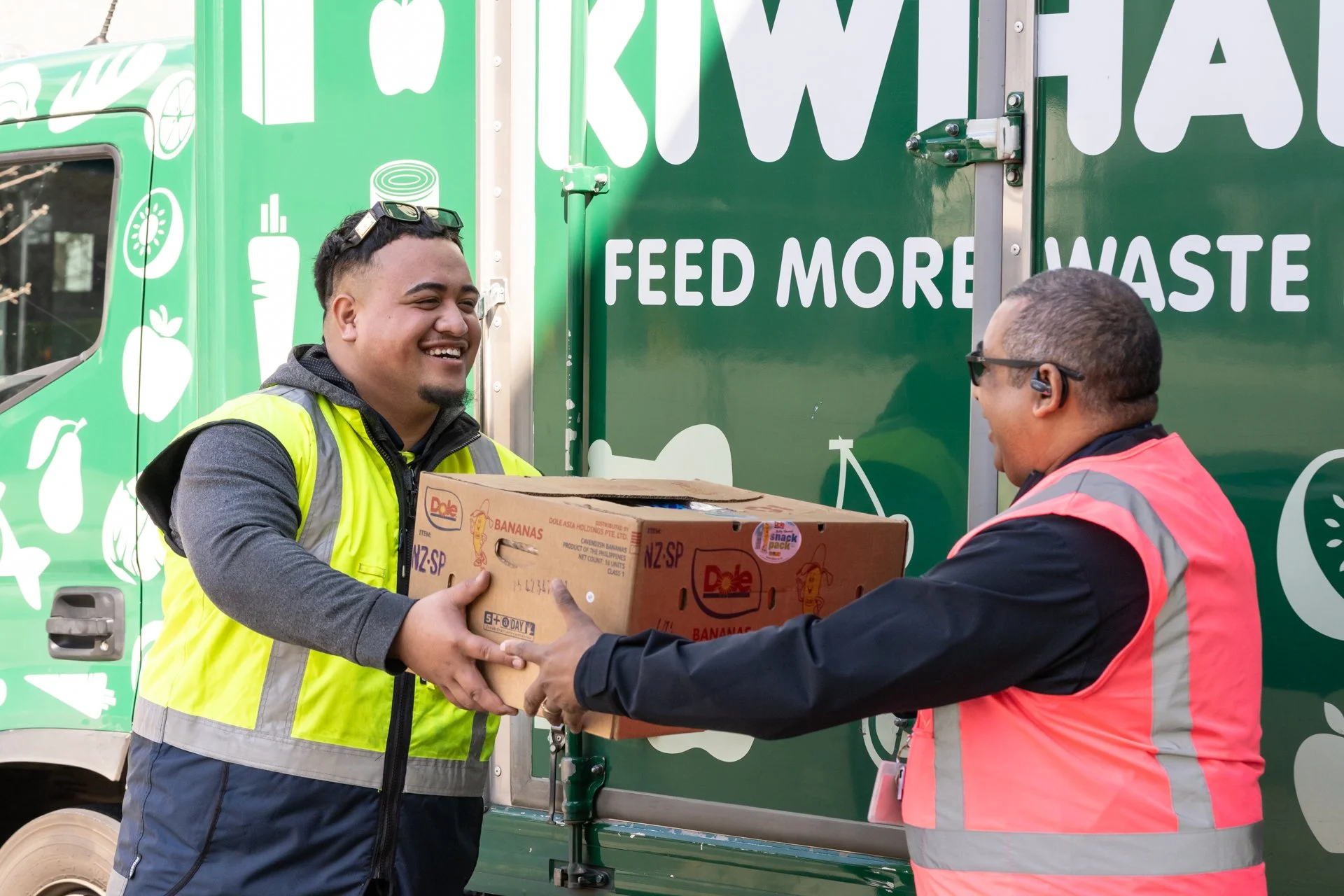 KiwiHarvest driver and volunteer stand smiling at the KiwiHarvest dept in Highbrook, handing a box of good surplus donated food.