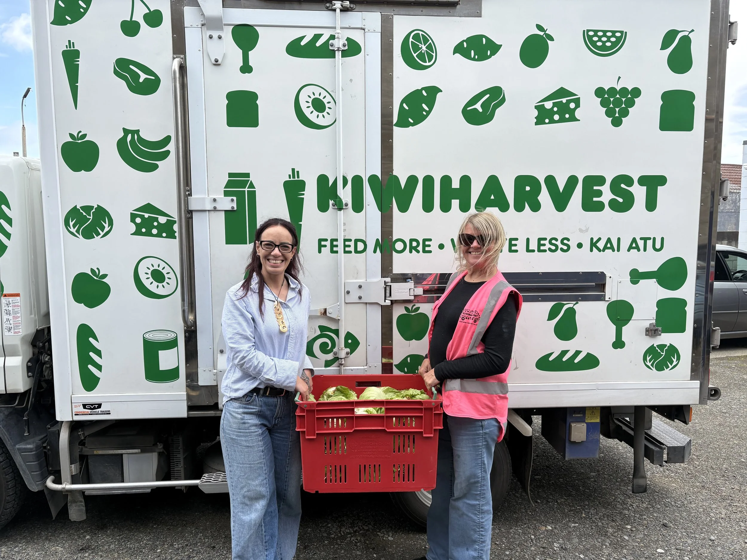Relationship manager from KiwiHarvest and staff member from a charitable partner stands holing a crate of fresh vegetables infront of the KiwiHarvest food rescue truck
