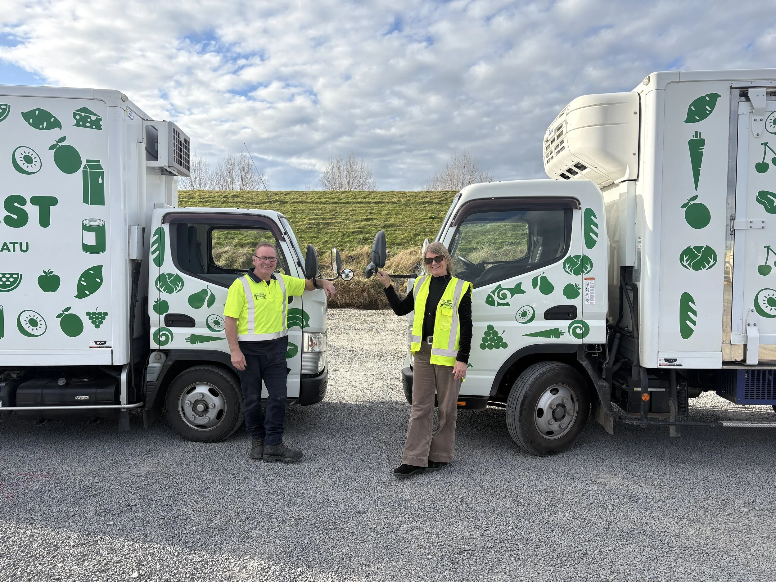 KiwiHarvest team members from Invercargill and Dunedin stand side by side smiling. Two KiwiHarvest trucks are in the background