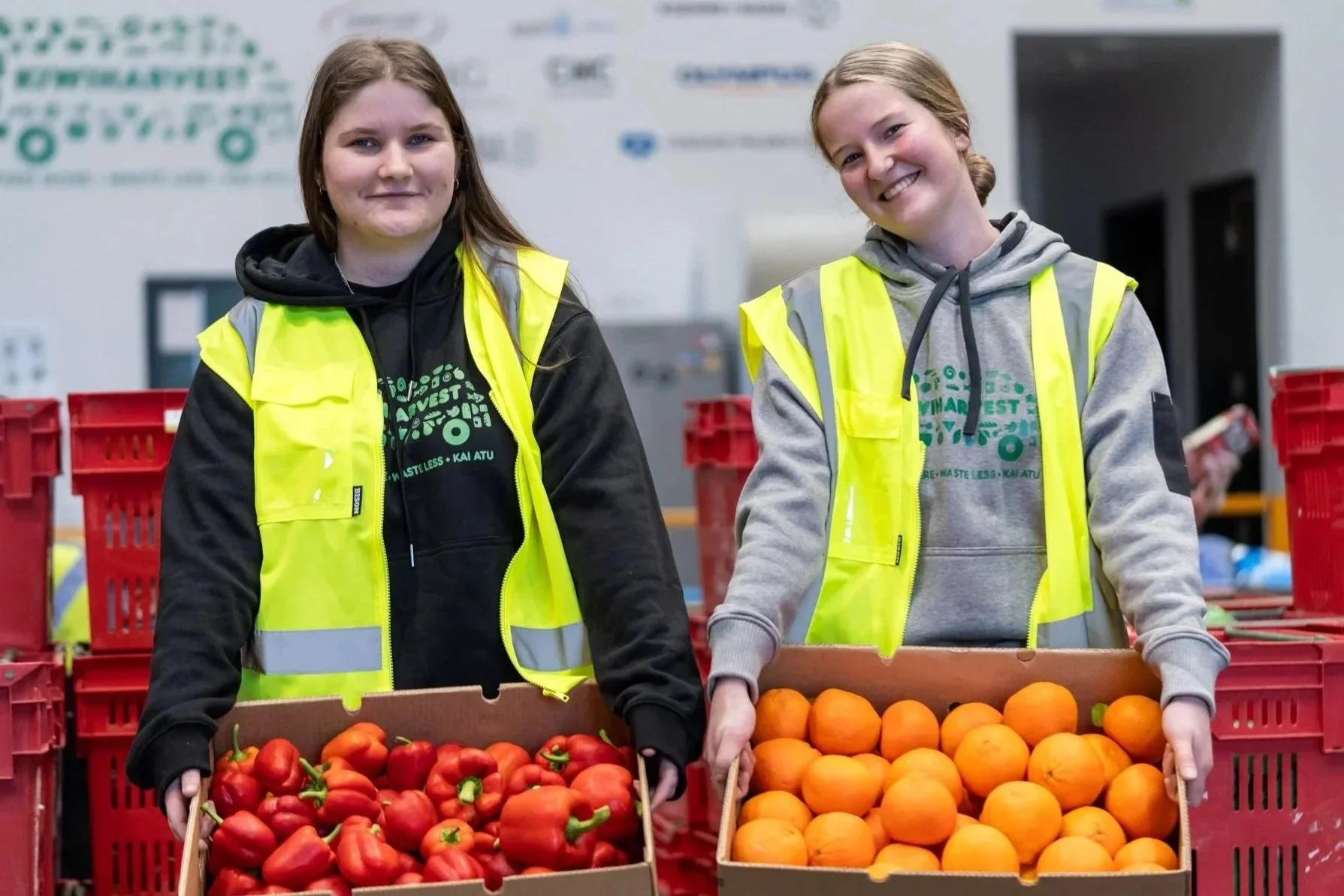 Two women from KiwiHarvest stand smiling with boxes of fresh donated produce 