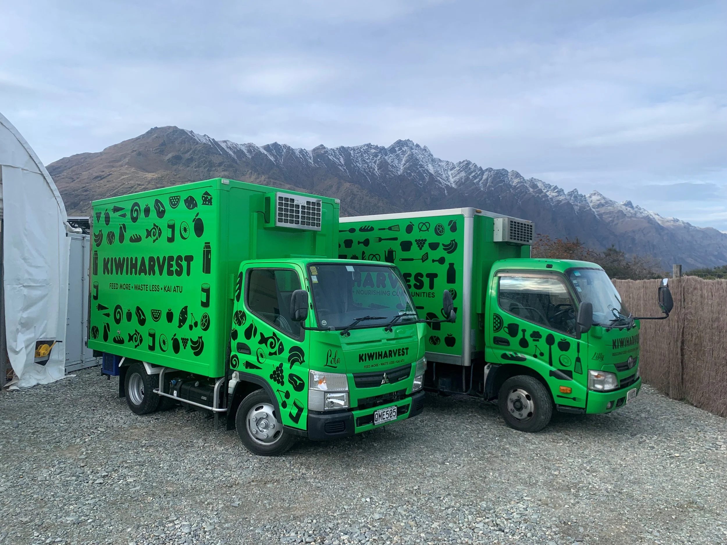 two Green KiwiHarvest trucks parked side by side. Snow top covered Queenstown mountain range in the background