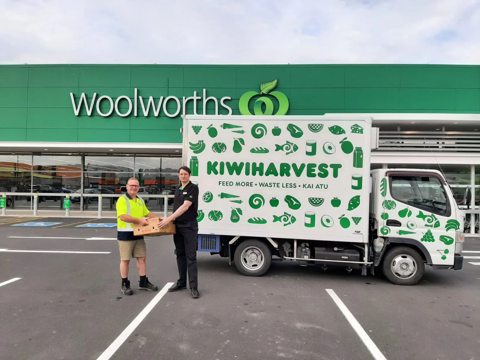 Woolworths store manager handing over a box of donated food to the Dunedin KiwiHarvest branch supervisor. They are standing outside of a Woolworthes supermarket next to a KiwiHarvest food rescue truck