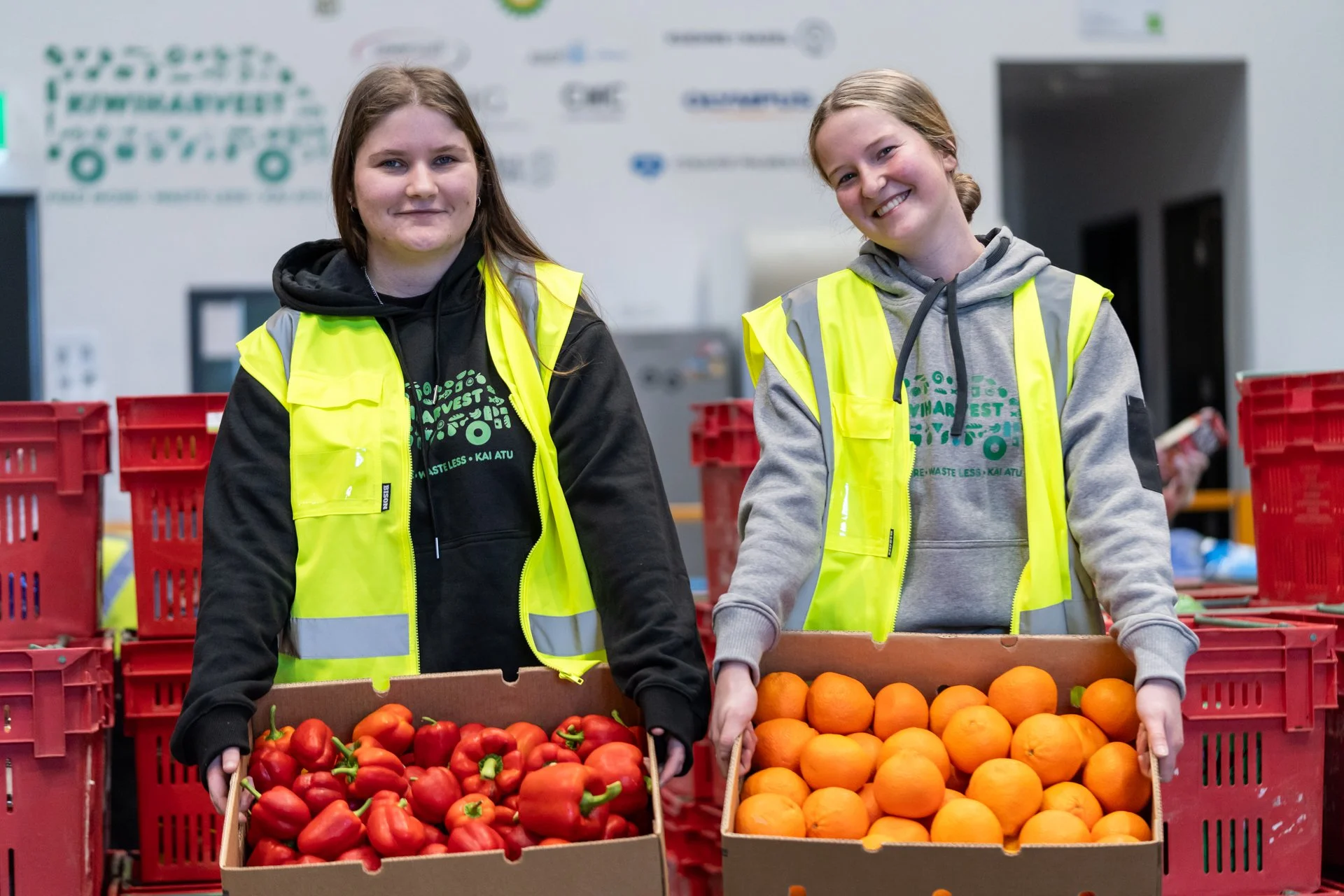 Two KiwiHarvest staff stand smiling holding boxes of fresh produce