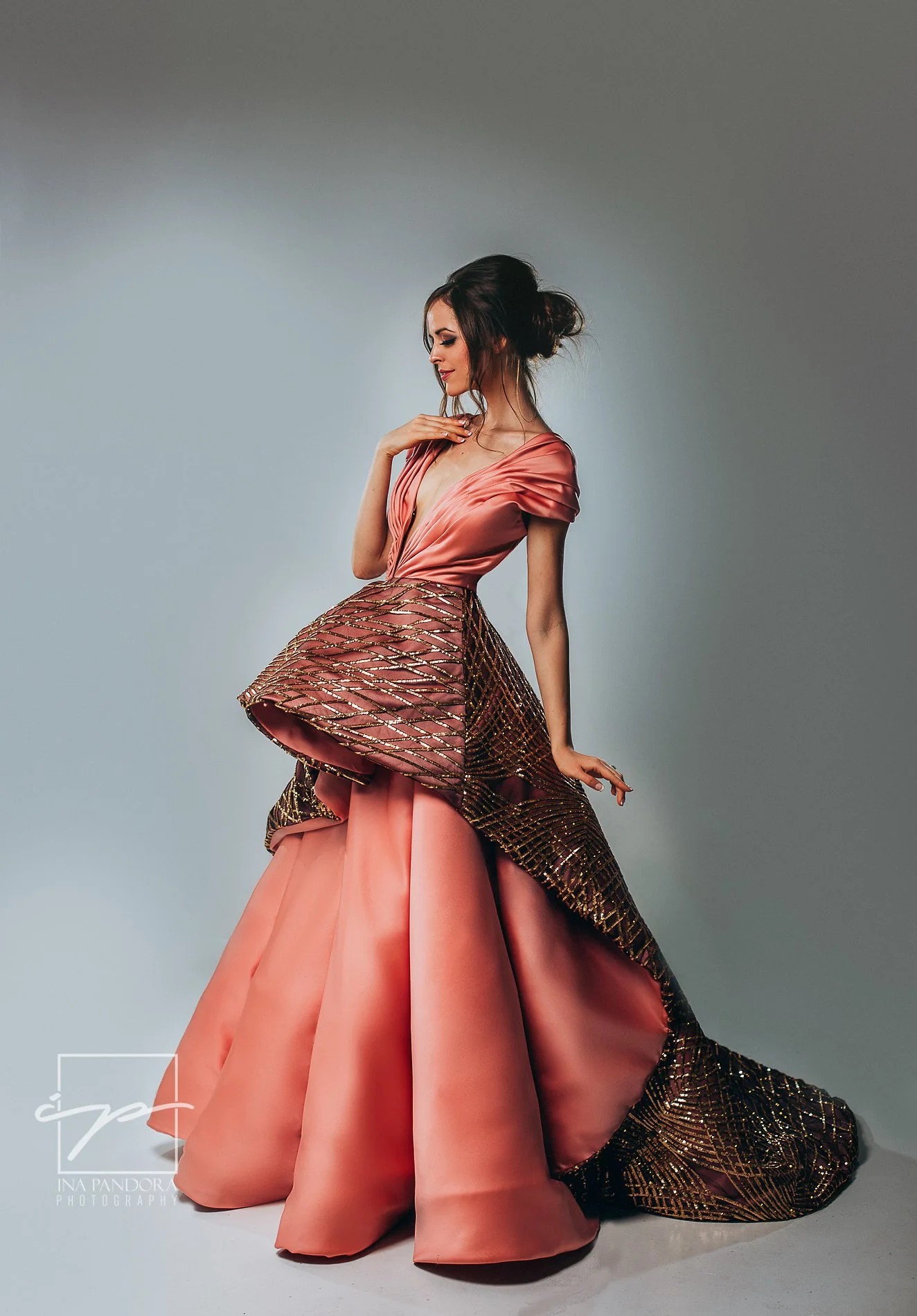 A woman in an elegant, layered peach and gold gown with puffy sleeves, intricate gold embroidery, and a flowing train, standing in a photography studio with a neutral backdrop.