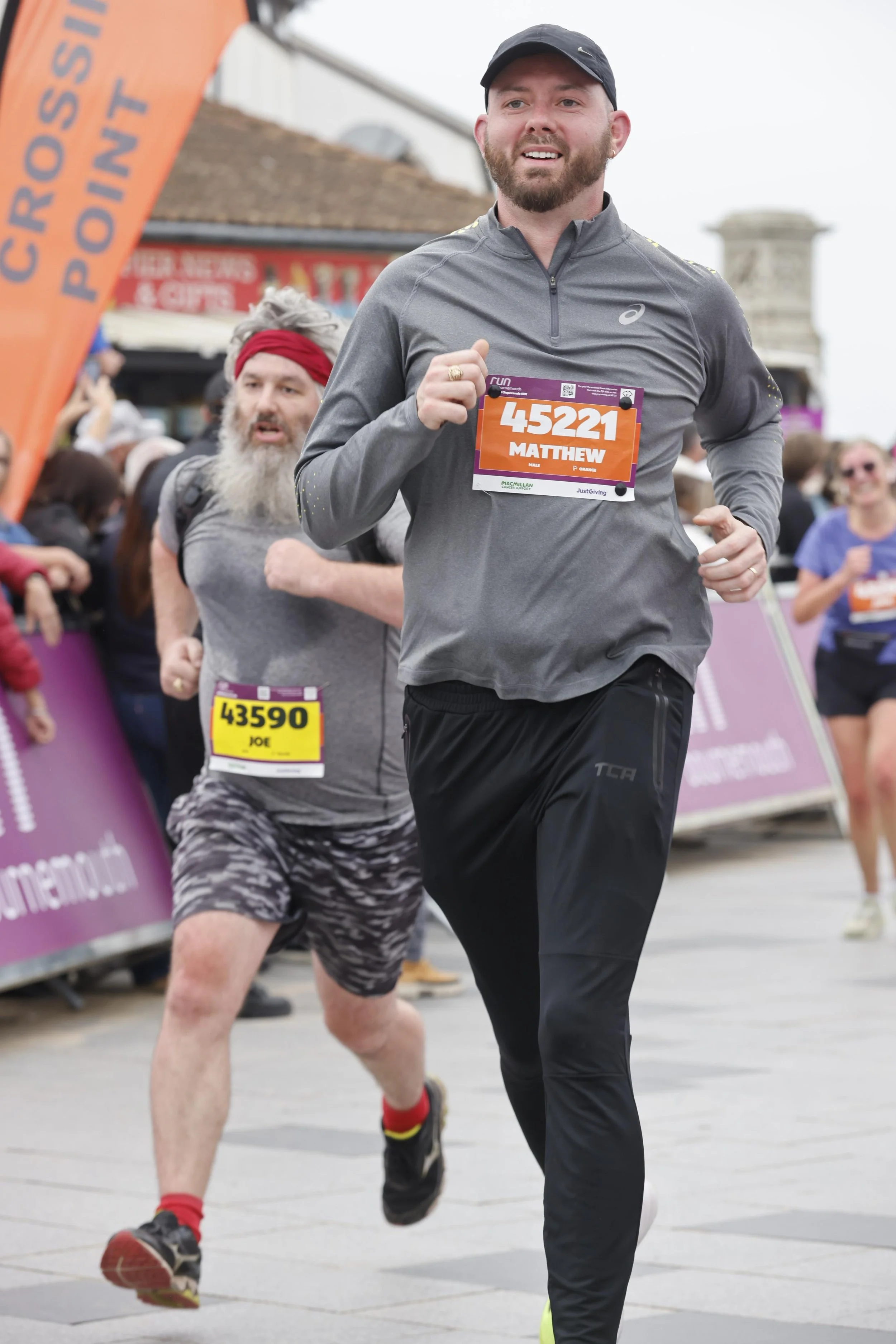 A man in a gray athletic jacket with a race bib reading 'Matthew' runs in a marathon, accompanied by another runner with a gray beard and red headband, among a crowd of spectators and cityscape.