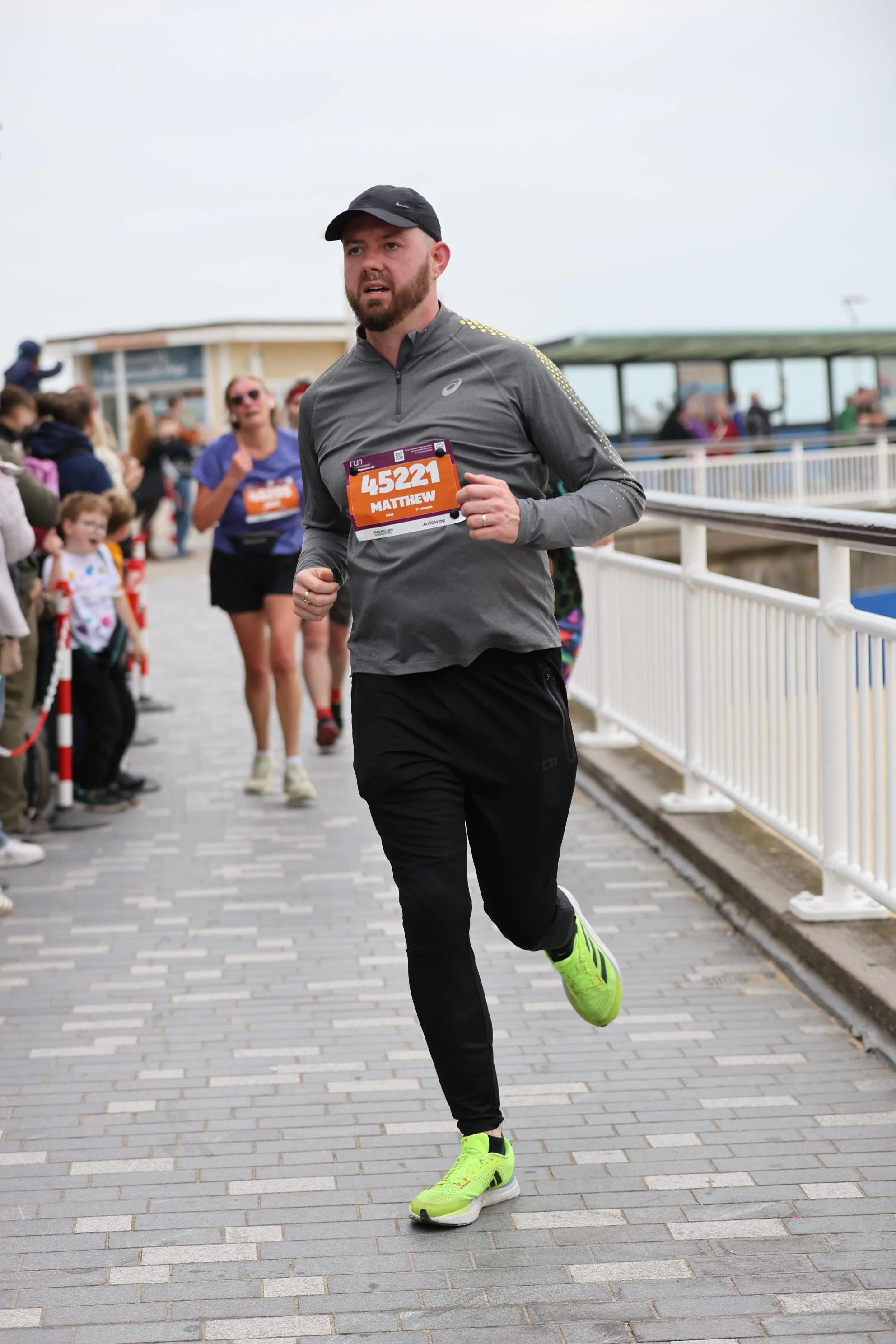 Man in running gear participating in a race, wearing a black cap, gray long-sleeve shirt, black pants, bright yellow running shoes, and a race bib with the number 45221 and name Matthew. He is running on a brick pathway near a white fence during daytime with spectators in the background.