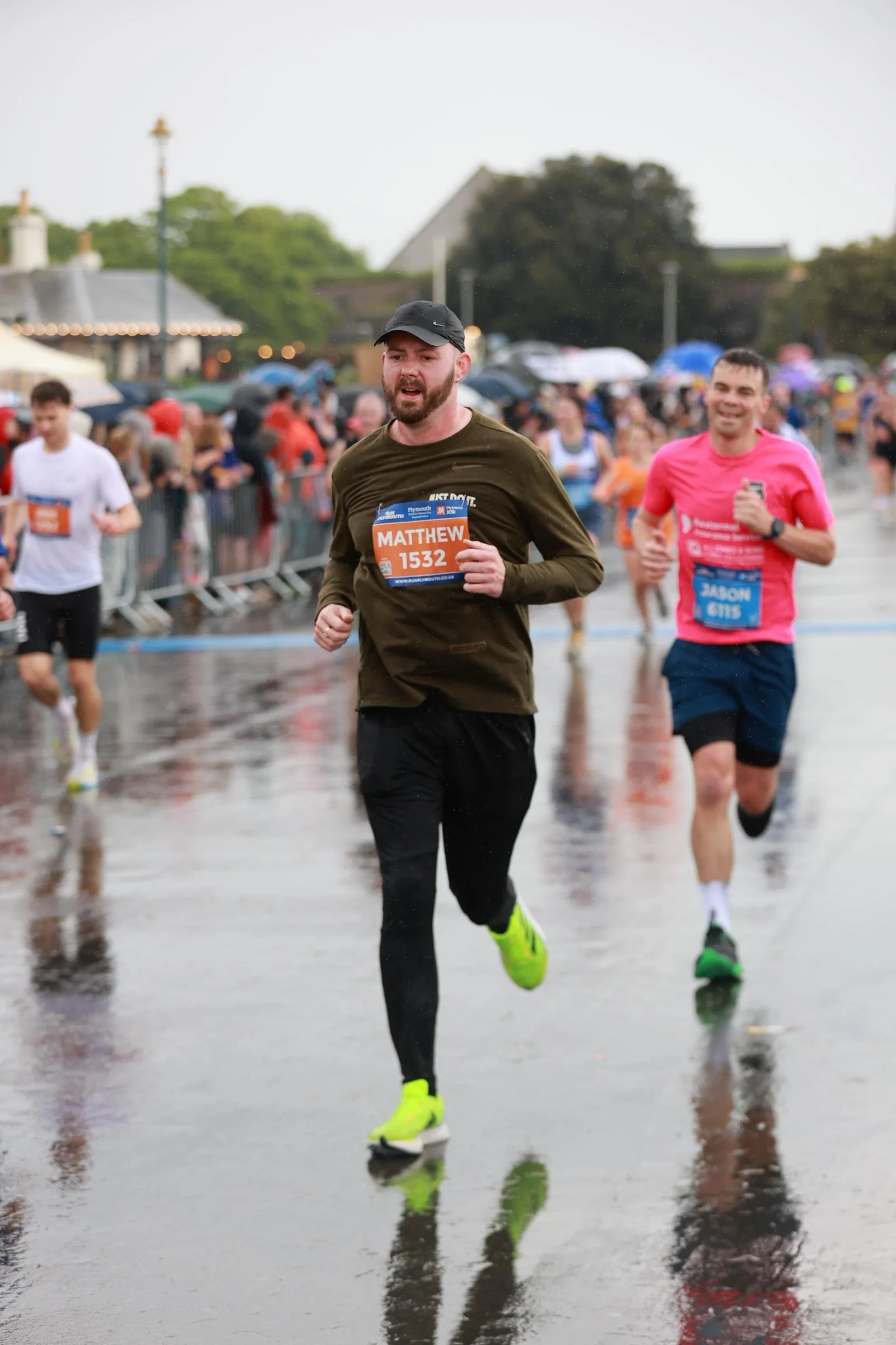 A man with a beard running in a marathon on a wet street, wearing a black cap, black pants, a green long-sleeve shirt, and neon yellow shoes. There are other runners and spectators holding umbrellas in the background.
