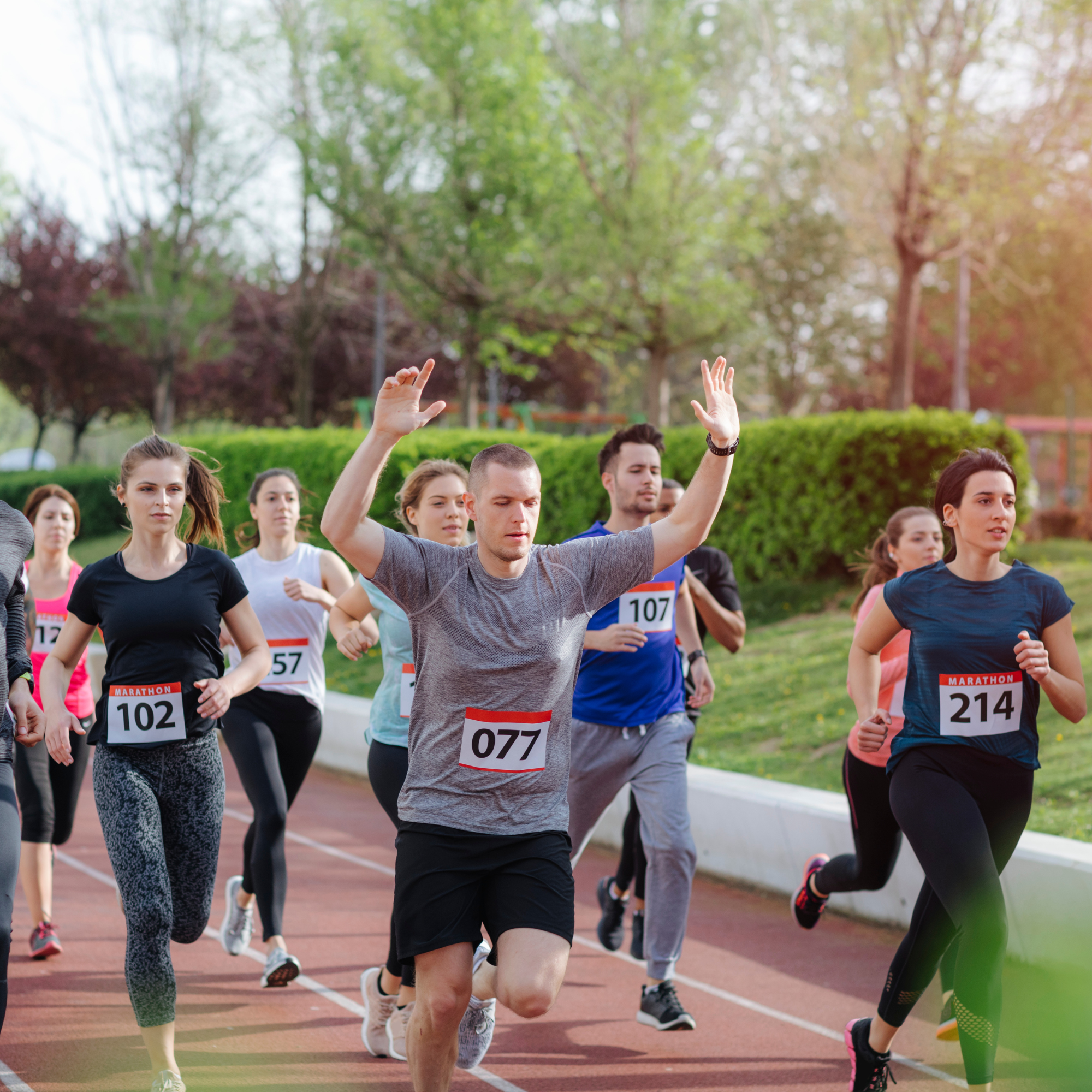 People participating in a marathon running event on a track in a park with trees in the background, raising their hands and wearing numbered bibs.